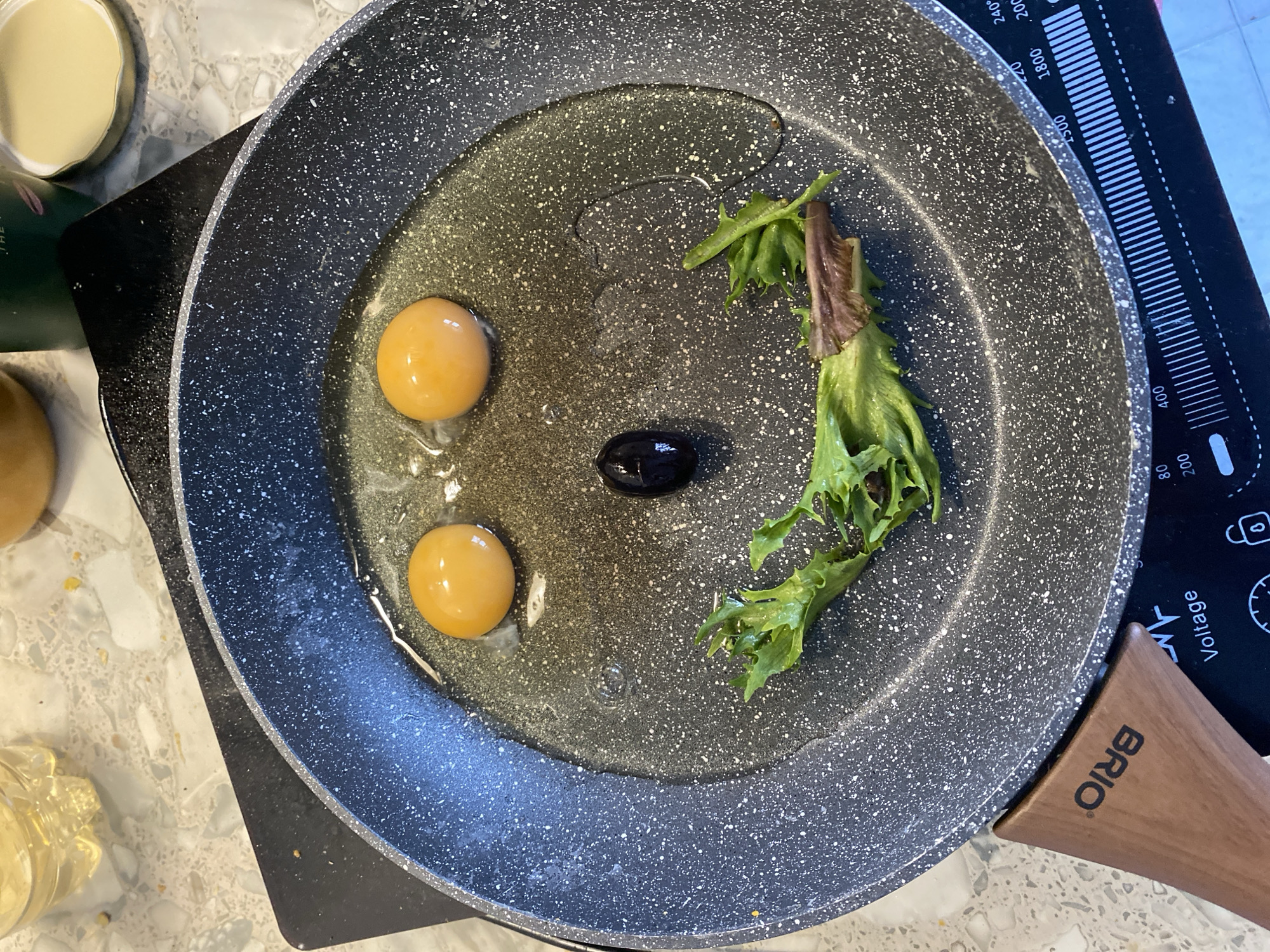 The scene depicts a non-stick frying pan positioned on a black induction cooktop within a kitchen setting in Pleven, Bulgaria. The pan contains cooking oil, two raw egg yolks, a single black olive, and a piece of green leaf lettuce, arranged to form a stylized smiley face. The egg yolks are located in the upper portion, the olive centrally below them, and the lettuce forms an arc in the lower portion of the pan. The pan has a dark gray, speckled interior and a brown, wooden-textured handle, partially visible at the lower left, marked with the brand "BRIO". The induction cooktop displays various power and temperature settings, including "Voltage" and numerical values such as "80", "200", "400", "1500", "1800", "240°", and "2000", indicating its operational status or controls. The countertop is a light-colored speckled surface. In the blurred background, a transparent plastic bottle is visible at the upper left, while a dark green container and a detached lid with a gold rim are present at the upper right. No individuals are present in the image.