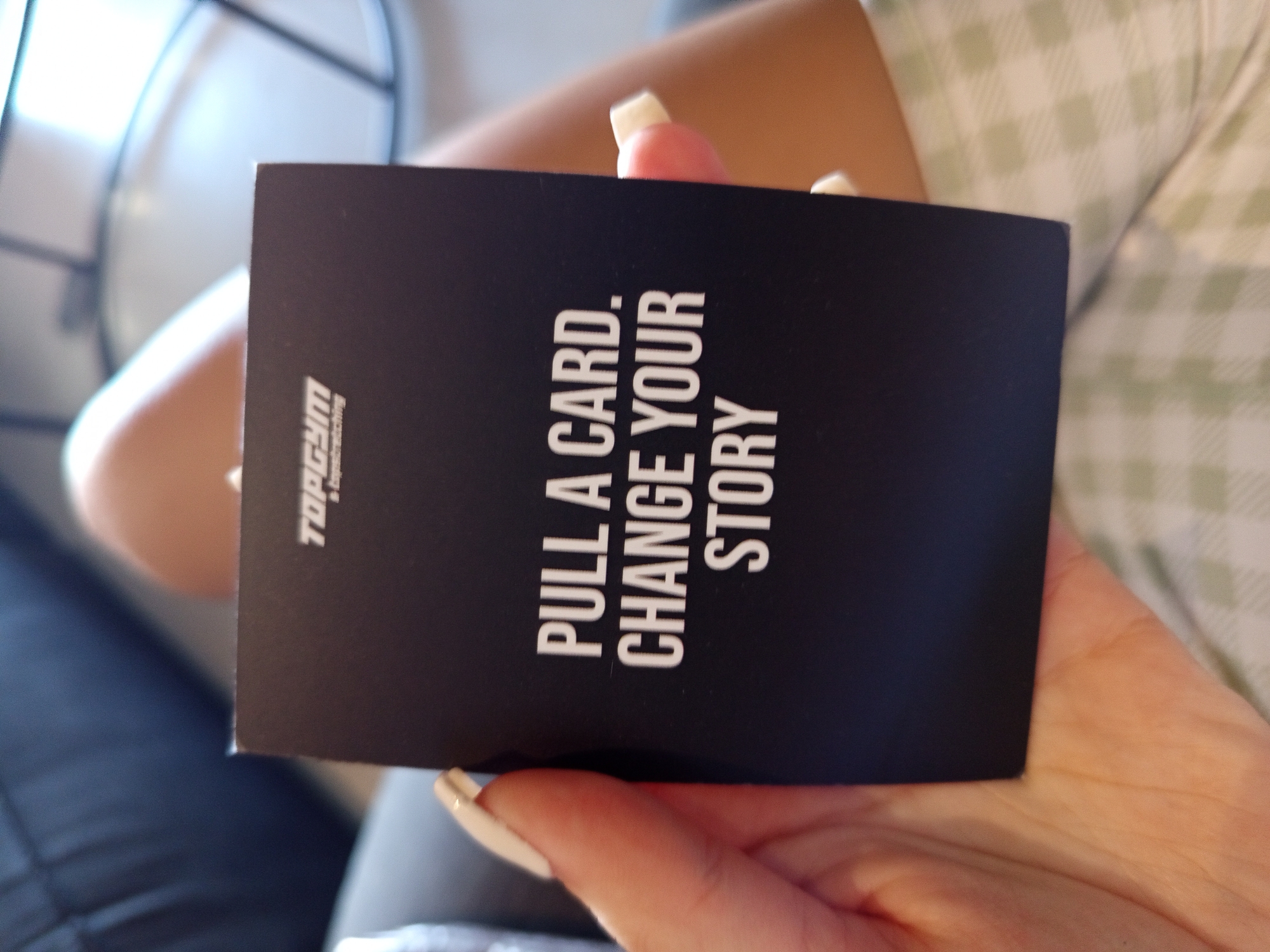 A person is holding a black card with white text that reads "PULL A CARD. CHANGE YOUR STORY" and the logo "TOPGYM by Topboxing" on the left side. The person's fingers with white polished nails are visible holding the card. The person's right thigh, covered in a white and green plaid garment, is visible in the background. A partially visible dark colored chair is in the background to the left. No information is available regarding the location in North Kuta, Indonesia from the given image.