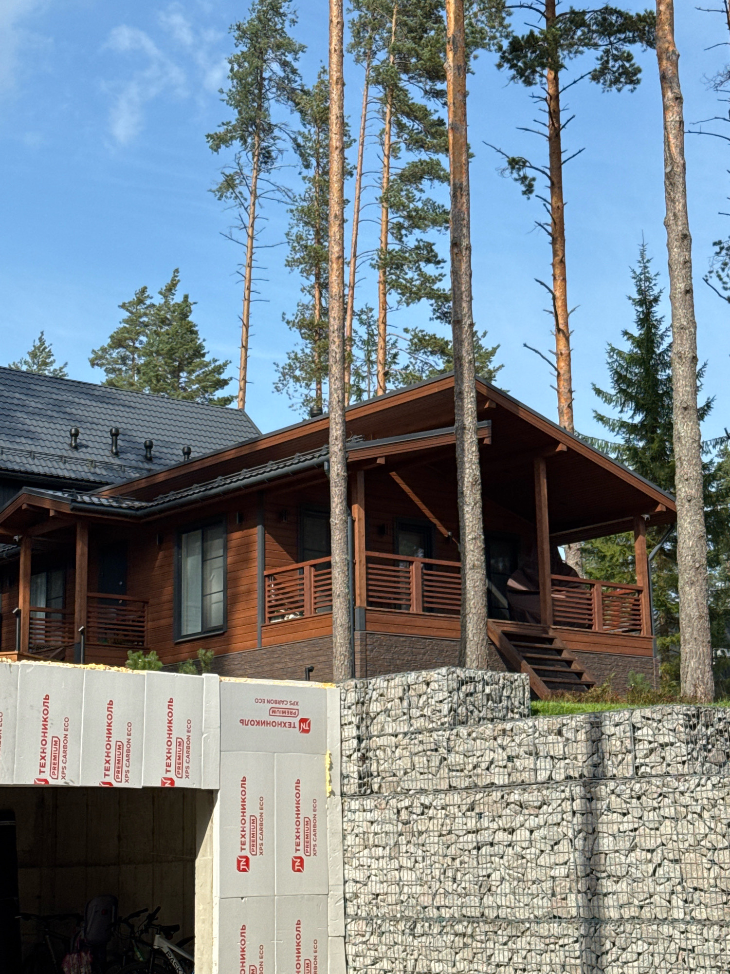 The image depicts a two-story wooden house with a dark gray tiled roof, located in the city of Mars, Russia. The house features a prominent covered porch with wooden railings and stairs leading up to it. Several tall pine trees are scattered around the property, with their trunks and branches visible against a clear blue sky. In the foreground, there are gabion walls constructed with wire cages filled with stones, forming a tiered landscaping feature. Partially visible at the bottom left of the frame is an open garage-like structure with bicycles stored inside. The construction of an adjacent building is evident, with white foam insulation boards stacked and bearing Russian text.