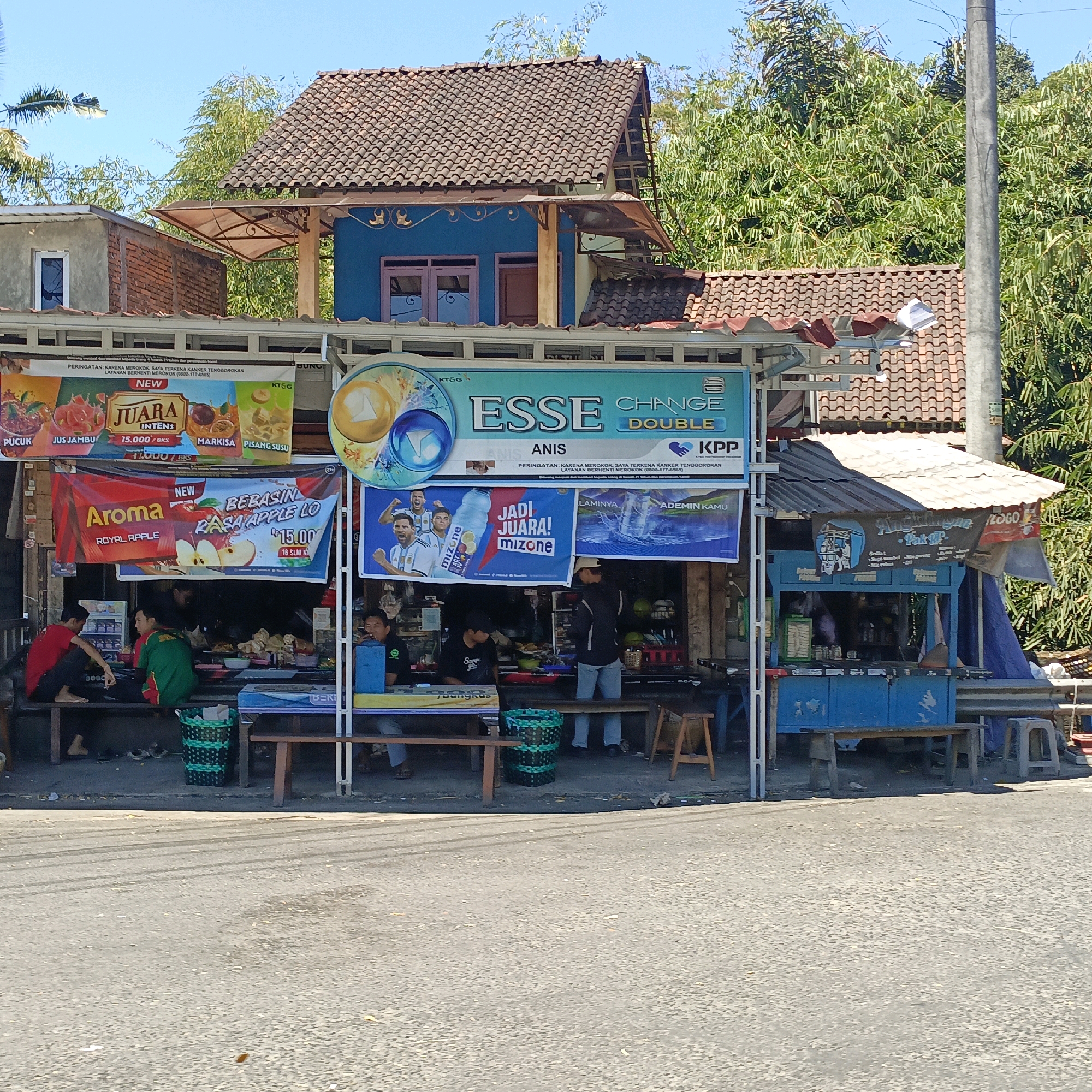 The image captures a street-level view of a small roadside establishment in Salatiga, Indonesia, likely a kiosk or food stall. The building features a multi-tiered roof structure with terracotta tiles. A bright blue section of the building has decorative gold motifs and a pink-framed window. Several signs are prominently displayed, advertising various products including "JUARA INTENS," "ESSE CHANGE DOUBLE," "Aroma ROYAL APPLE," and "MIZONE." One sign includes an image of soccer players, likely referencing a promotional campaign.
In the foreground, people are seated on a wooden bench, some appearing to be customers and others potentially vendors. A green woven basket is placed on the ground. Further to the right, another kiosk-like structure is visible, painted blue, with a sign that reads "Angkringan Pak TIP." Various items, likely food and beverages, are displayed on shelves and tables. The overall scene suggests a bustling, informal commercial area with a focus on refreshment sales. The street in front appears to be asphalt. The background is dominated by lush green foliage, indicating a location with ample vegetation. A concrete utility pole stands to the right of the frame. The sky is clear and blue, suggesting a sunny day.
