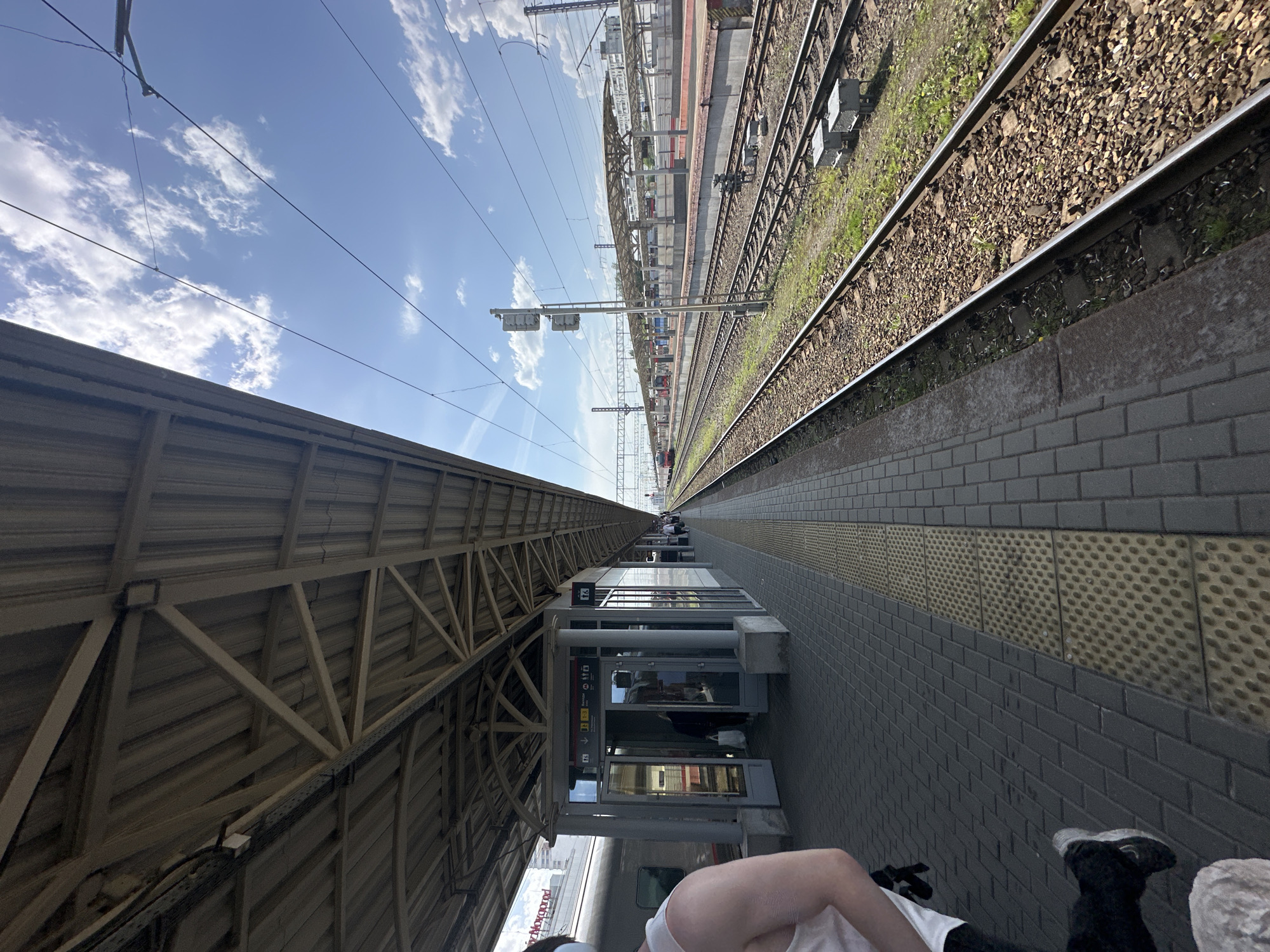 The image depicts a railway platform in Nizhny Novgorod, Russia. The perspective is from a low angle, looking along the length of the platform towards the horizon. The platform is paved with gray bricks, with yellow textured sections near the edge. A metal roof structure covers the platform. A train is partially visible on the left. Railway tracks run parallel to the platform on the right. The sky is blue with scattered clouds. Overhead power lines are visible. A person's torso and legs are visible in the bottom right corner of the image. There are informational signs located on the buildings adjacent to the train platform. There are other buildings alongside the train tracks.