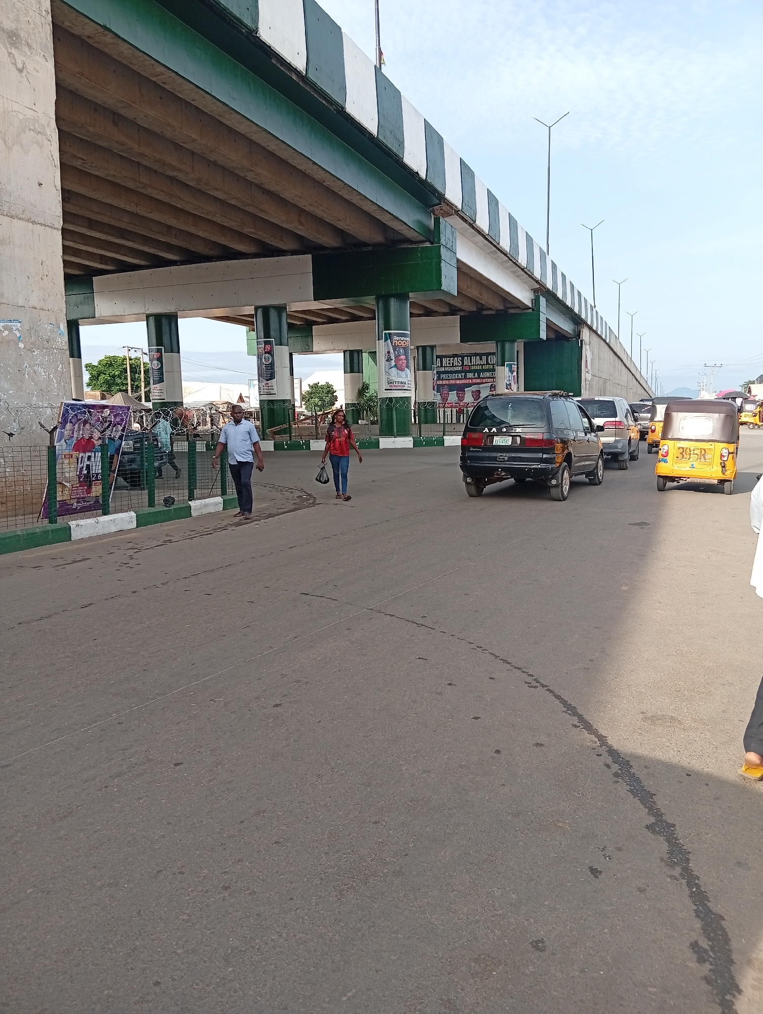 The image depicts a street scene in Jalingo, Nigeria, under a large overpass. A man in a blue shirt and dark trousers walks on the asphalt road, holding a cane. A woman in a red t-shirt and blue jeans walks in the same direction, carrying a blue bag. Several vehicles are visible on the road, including a black SUV, a white car, and two yellow auto-rickshaws. The overpass is made of concrete with green and white striped railings. Billboards advertising political campaigns are posted along the side of the road. The sky is partially cloudy.