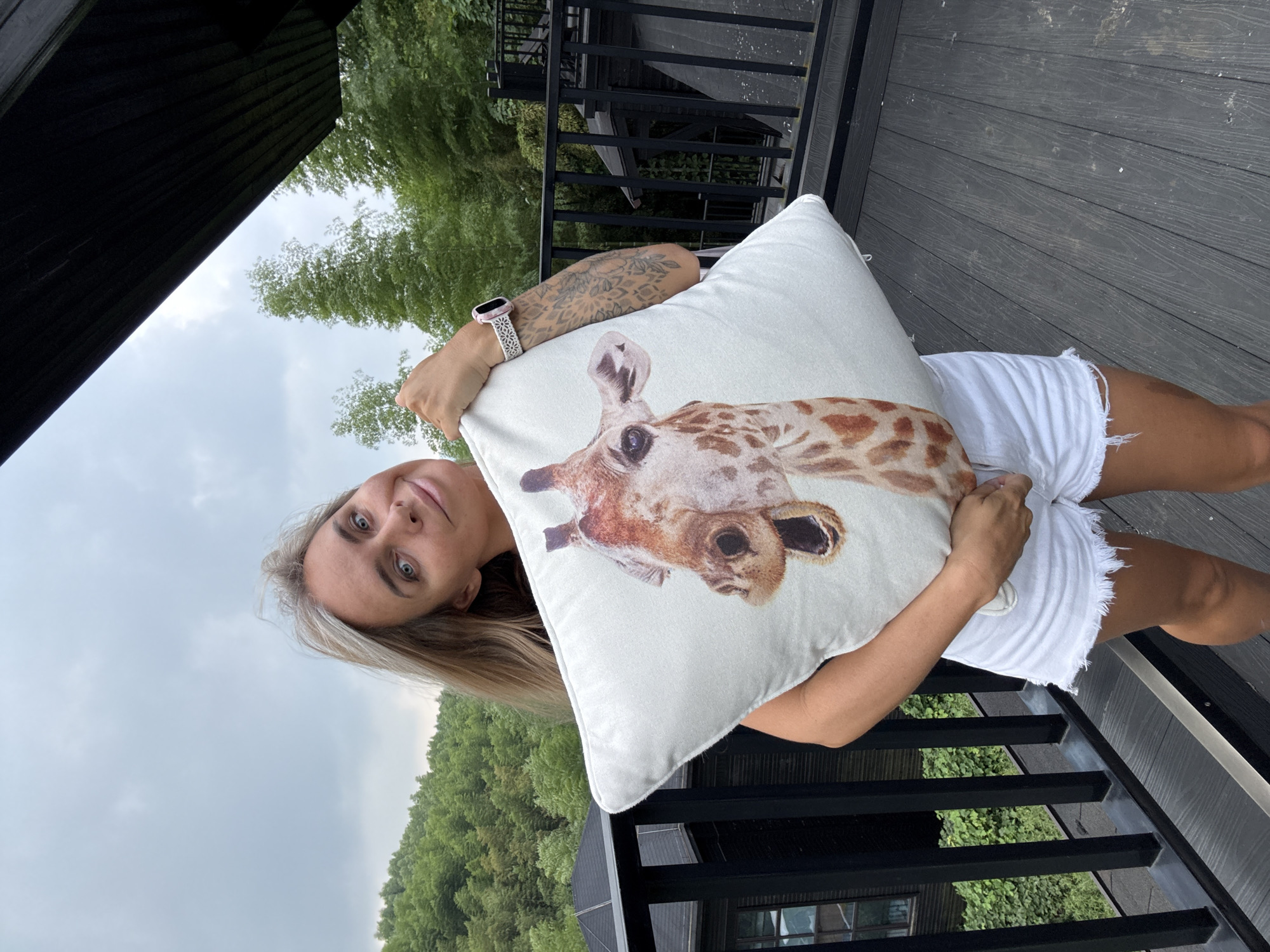 A woman with blonde hair and blue eyes, wearing white denim shorts and a black top, stands on a wooden deck holding a white pillow with a giraffe's face printed on it. She has tattoos on her left arm and is wearing a smartwatch on her left wrist. The deck has a black railing, and in the background, there are green trees and a cloudy sky. A portion of a dark, sloped roof structure is visible above and to the right. The overall scene suggests an outdoor setting, possibly a balcony or porch, with a natural, hilly landscape in the distance.