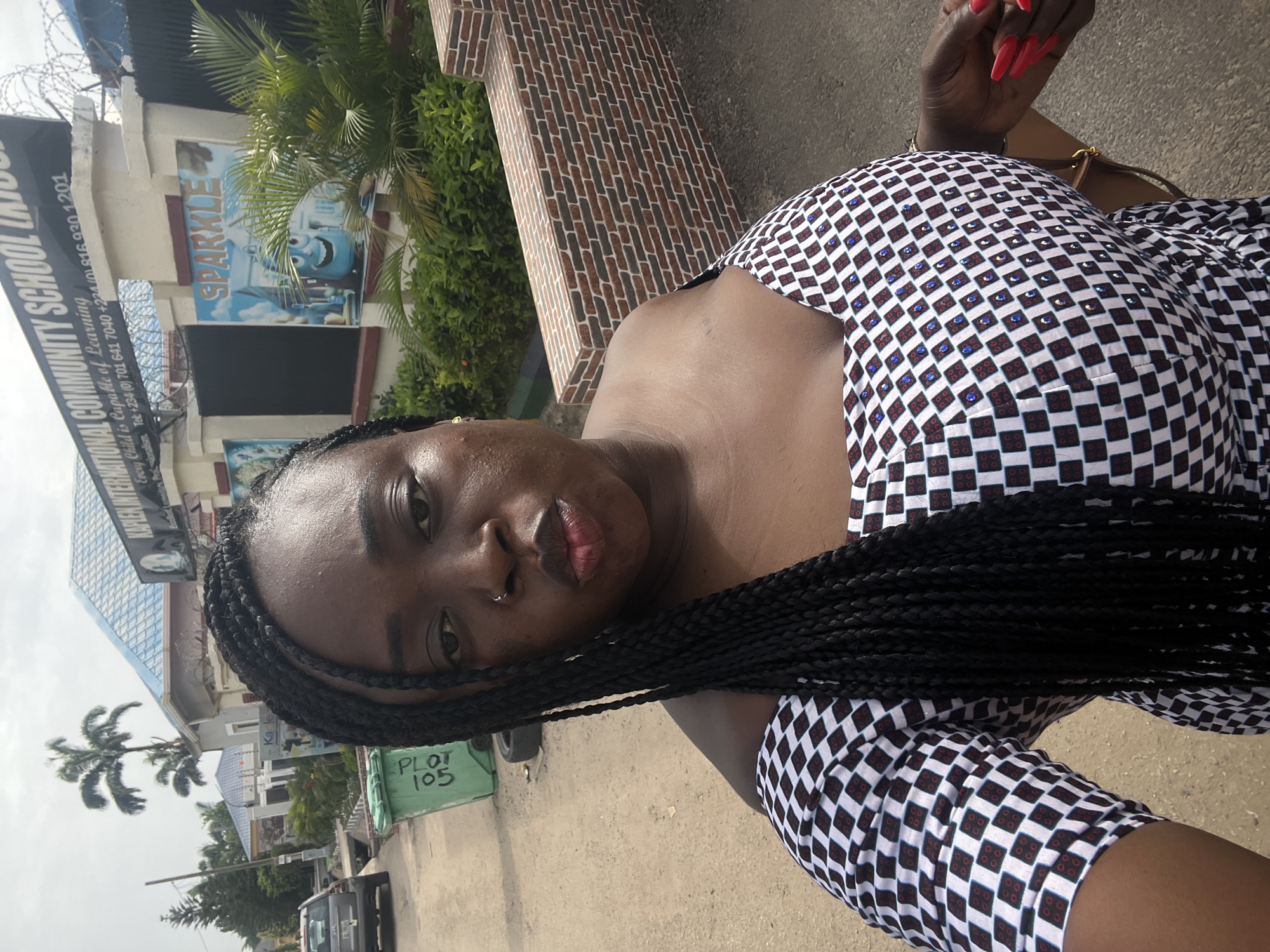 A woman is shown in a selfie taken in front of Maple International Community School in Abuja, Nigeria. The school's signboard is visible, listing contact numbers. The building behind her has a "Sparkle" sign and a brick facade. A palm tree and another building are visible in the distance. The woman is wearing a black and white patterned dress with small embellishments and has her hair in braids. She is wearing a gold-colored bracelet and has red nail polish. The green painted sign "PLOT 105" is seen on a low wall next to the street.