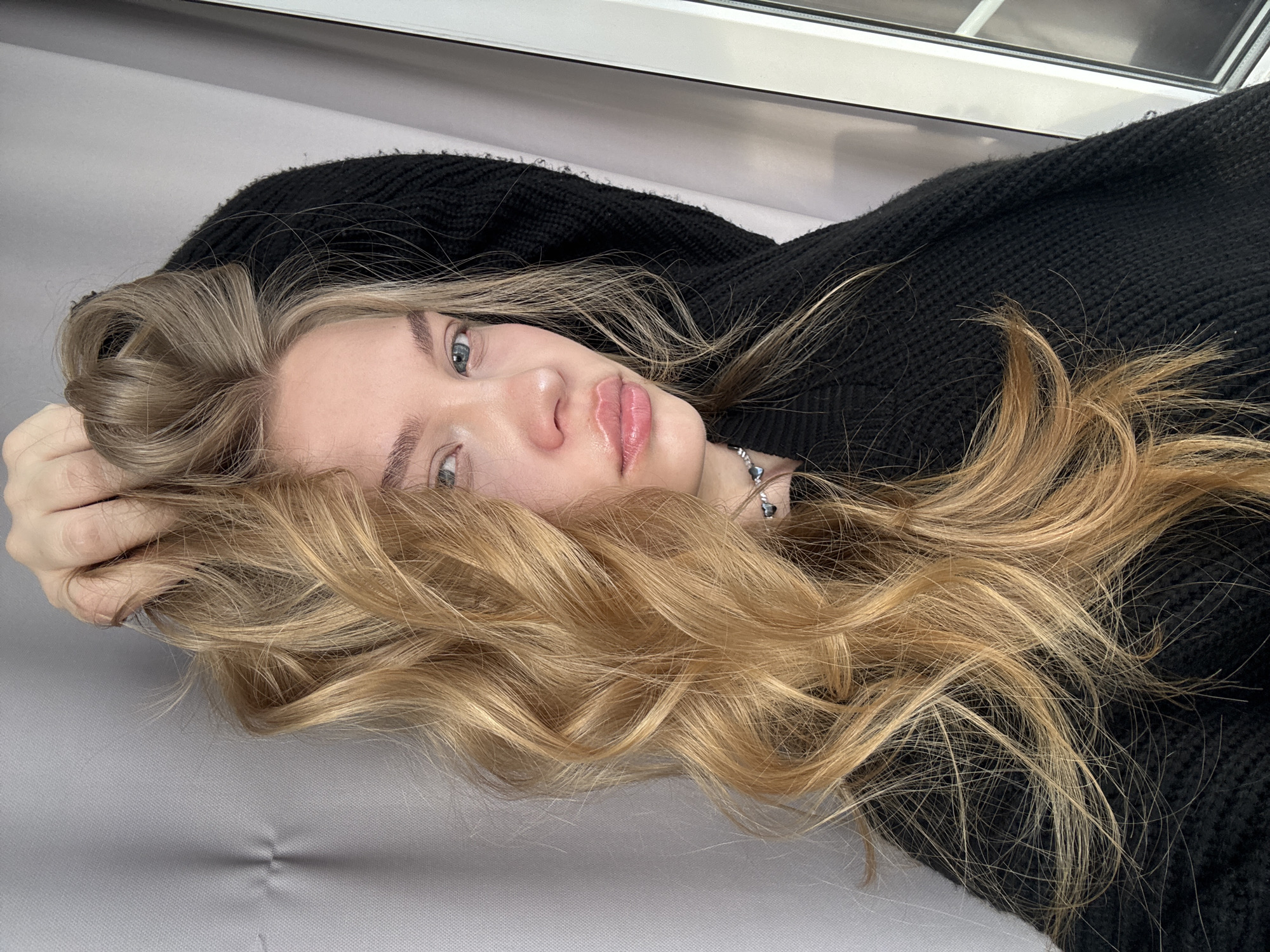 A young woman with light blonde hair is lying on a gray surface, likely a couch or bed. She is wearing a black knit sweater. She is looking directly at the camera. A silver necklace is visible around her neck. The location is possibly Chaikovsky, Russia, based on the provided request, but there is no confirming evidence within the image itself. The environment is relatively clean and simple. There is a window frame visible in the background.