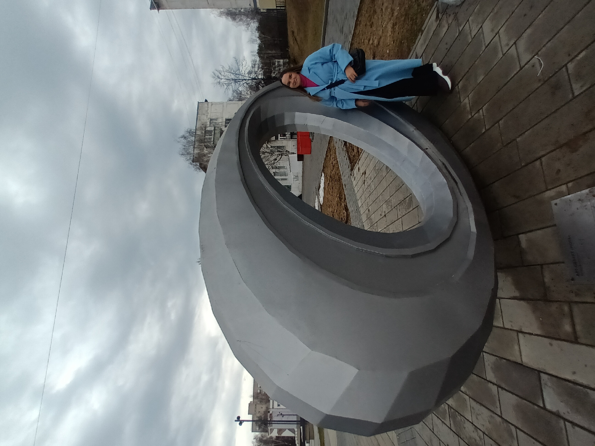 The image shows a woman leaning against a large, gray, toroidal sculpture outdoors. She is wearing a light blue coat over a pink top, black pants, and white sneakers. She has a black bag hanging on her shoulder. The sculpture is the dominant feature, with a multifaceted, rounded shape. The surrounding environment includes a paved area, patches of brown grass, and a few leafless trees. In the background, there are buildings, some appearing to be residential apartments. The sky is overcast with gray clouds. Based on the search results, this location is in the city of Chaikovsky, Perm Krai, Russia.