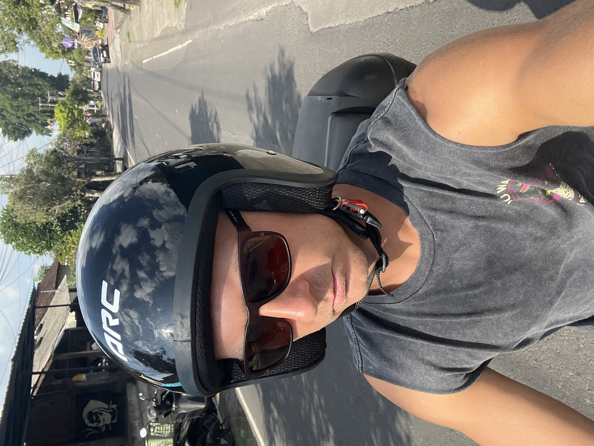 A man wearing a black helmet with white lettering that reads "ARC" and dark sunglasses is riding a scooter on a paved road in Sukawati, Indonesia. He is also wearing a dark grey sleeveless shirt. The road is lined with buildings, trees, and other scooters. The sky is blue with some clouds. The man's face is visible, and he appears to be looking forward. The image is taken from his perspective, likely a selfie.