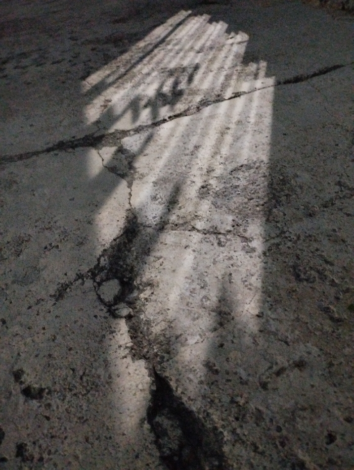 The image displays a weathered, grey concrete surface, possibly a floor or ground, located in the city of Guacara, Venezuela. The concrete is heavily cracked, with dark fissures of varying widths running across the frame, notably a large, irregular crack extending from the bottom center upwards. A rectangular pattern of parallel light stripes, consistent with sunlight filtering through an overhead grate or slatted opening, is cast diagonally across the surface from the upper right to the lower left. The lighting is dim, highlighting the textured, granular appearance of the concrete and the pronounced shadows. No individuals, distinct objects other than the concrete, or actions are present.