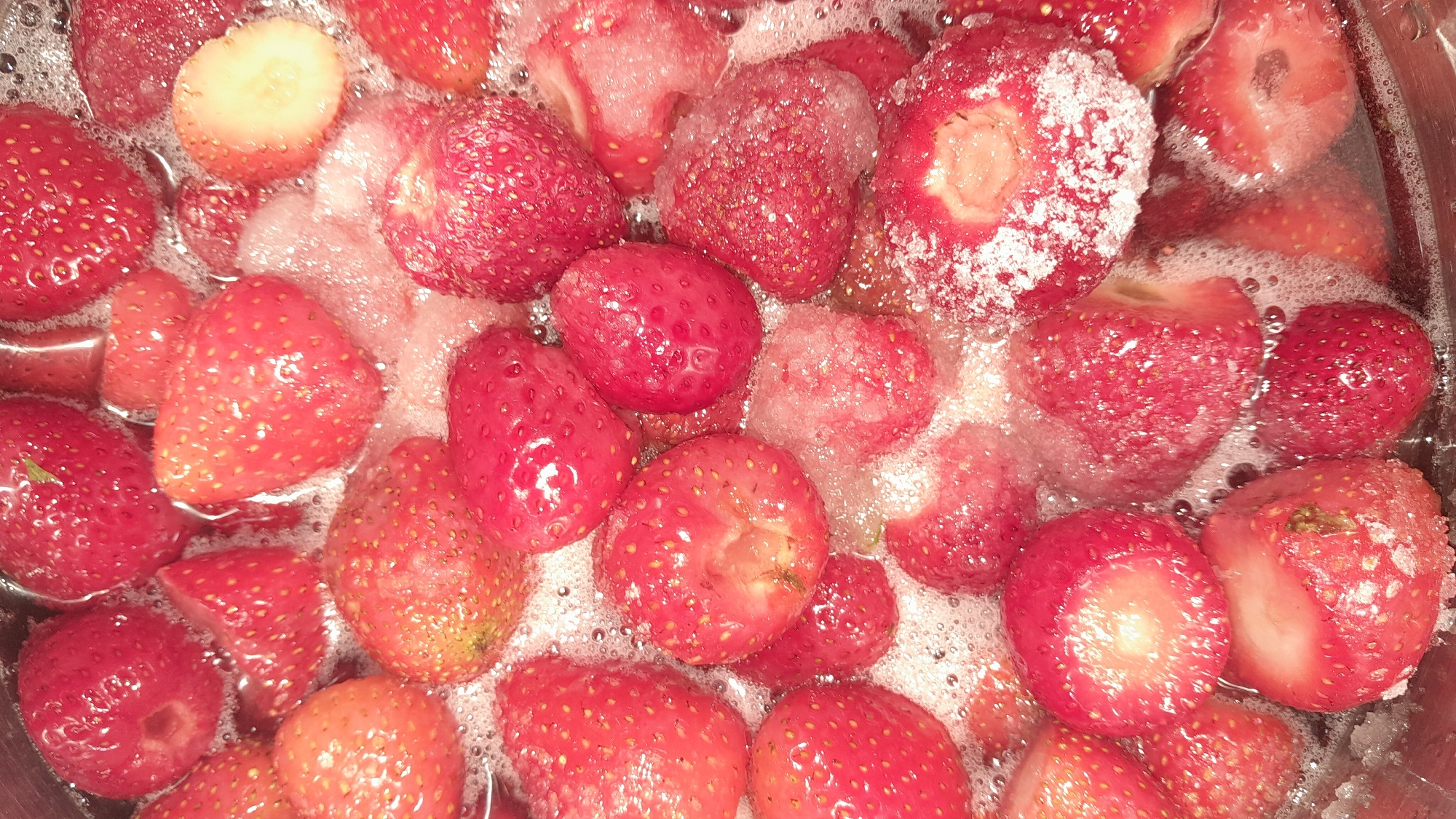 A close-up, top-down shot shows a pot of strawberries submerged in liquid and sugar, with a layer of foamy bubbles on the surface. The strawberries are a mix of whole and halved fruits, with their vibrant red color visible beneath the bubbly mixture. Some strawberries appear to be glistening, suggesting they are being cooked or preserved. The scene is likely indoors, possibly in a kitchen, given the context of cooking or preparing food. There are no people or animals visible in the frame. The lighting appears to be diffused, creating a soft glow on the fruits. The overall impression is one of a food preparation process, likely for making jam or a similar preserve. The location is not specified as being in any city.