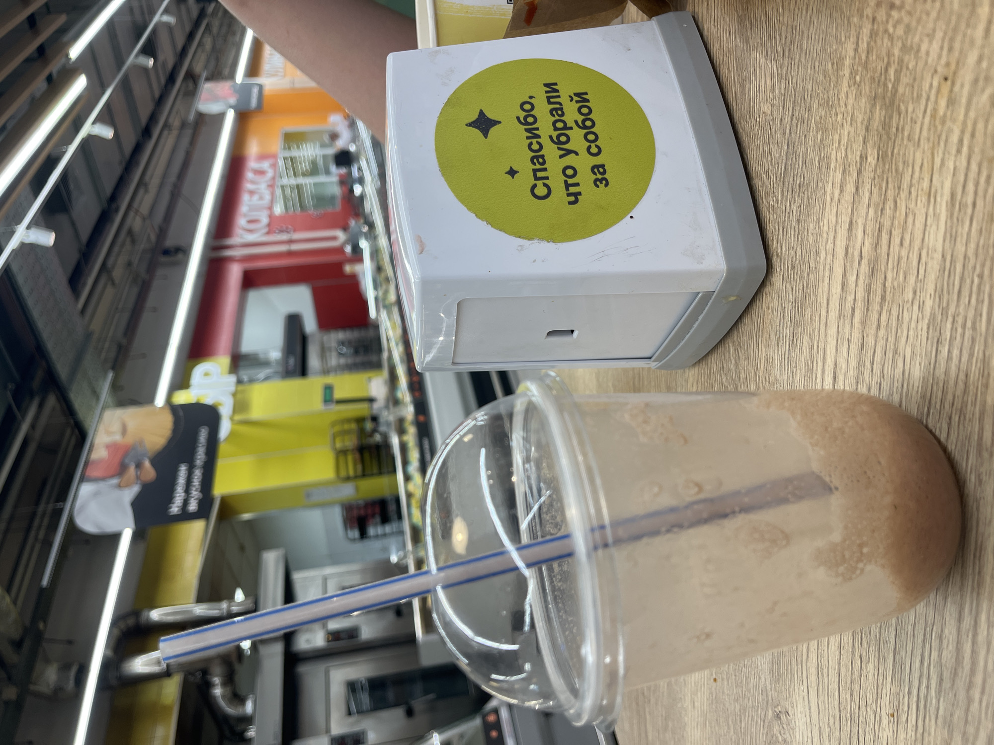 The image depicts a close-up view of a table in what appears to be a cafe or food service area within a supermarket in Volzhsky, Russia. In the foreground, a partially consumed beverage in a clear plastic cup with a lid and straw sits on a wooden table. To the right of the cup is a white napkin dispenser with a green circular sticker. The sticker has a black star-like symbol at the top and Cyrillic text that reads "Спасибо, что убрали за собой" (Thank you for cleaning up after yourself). The background reveals the interior of a commercial establishment, possibly a supermarket aisle, with bright lighting, shelving units, and signage. Notably, a sign with "КОЛБАСА" (Sausage) is visible in the background, suggesting a food retail environment. The overall scene is a casual snapshot of a personal moment, likely taken by a customer.
