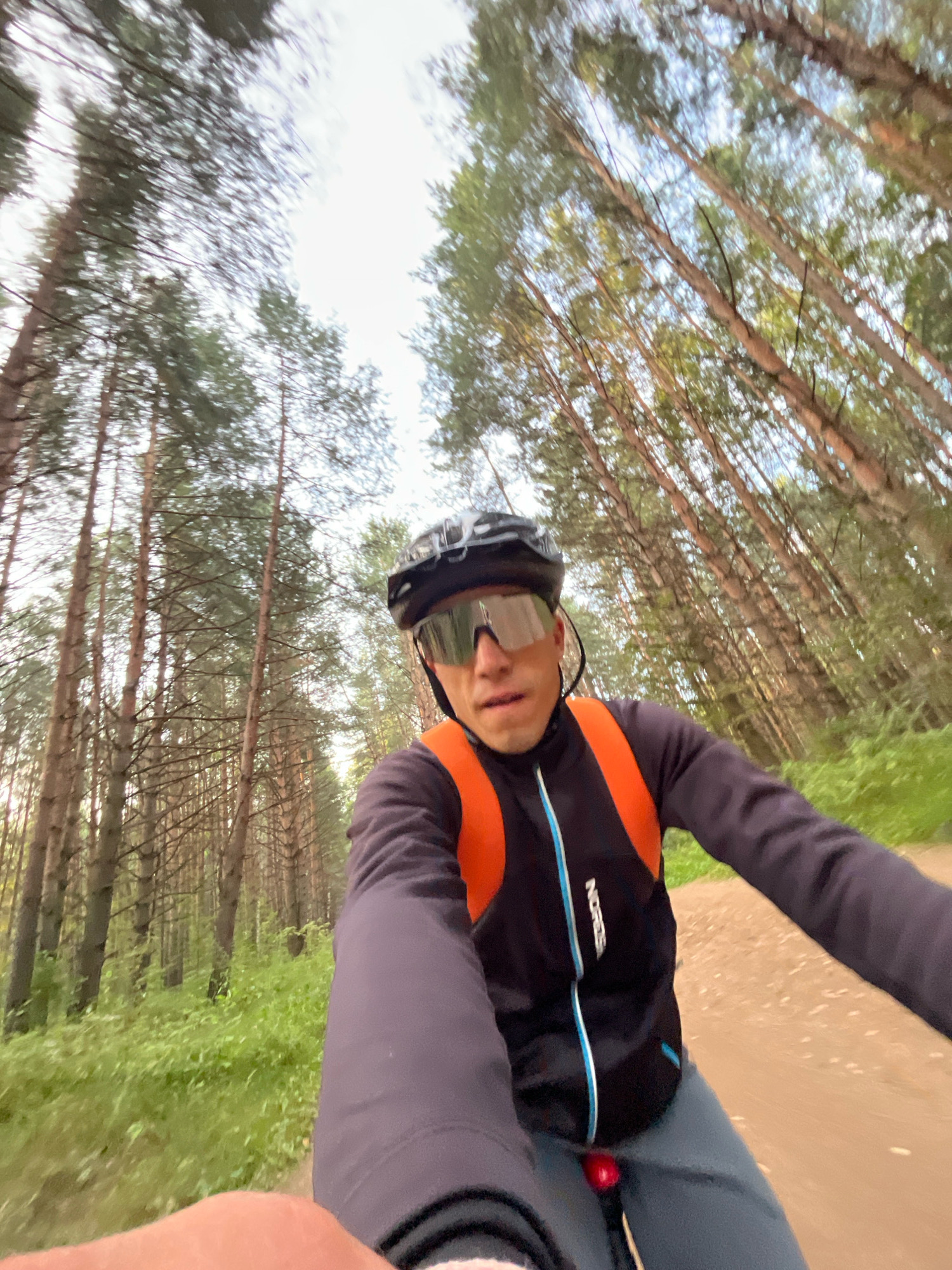 A first-person perspective selfie video captures a man cycling through a dense forest. The man, wearing a black helmet, sunglasses, and a black and orange cycling jacket, is the primary subject. He is positioned in the foreground, with the forest and sky blurred in the background, indicating rapid motion. The trees, predominantly tall and thin pines, line a dirt path. The ground is covered in green grass and foliage. The lighting suggests it is daytime, possibly late afternoon or early morning, with soft sunlight filtering through the trees. The scene is likely a trail in or near the city of Chaikovsky, Russia, a location known for its natural surroundings and recreational outdoor activities.