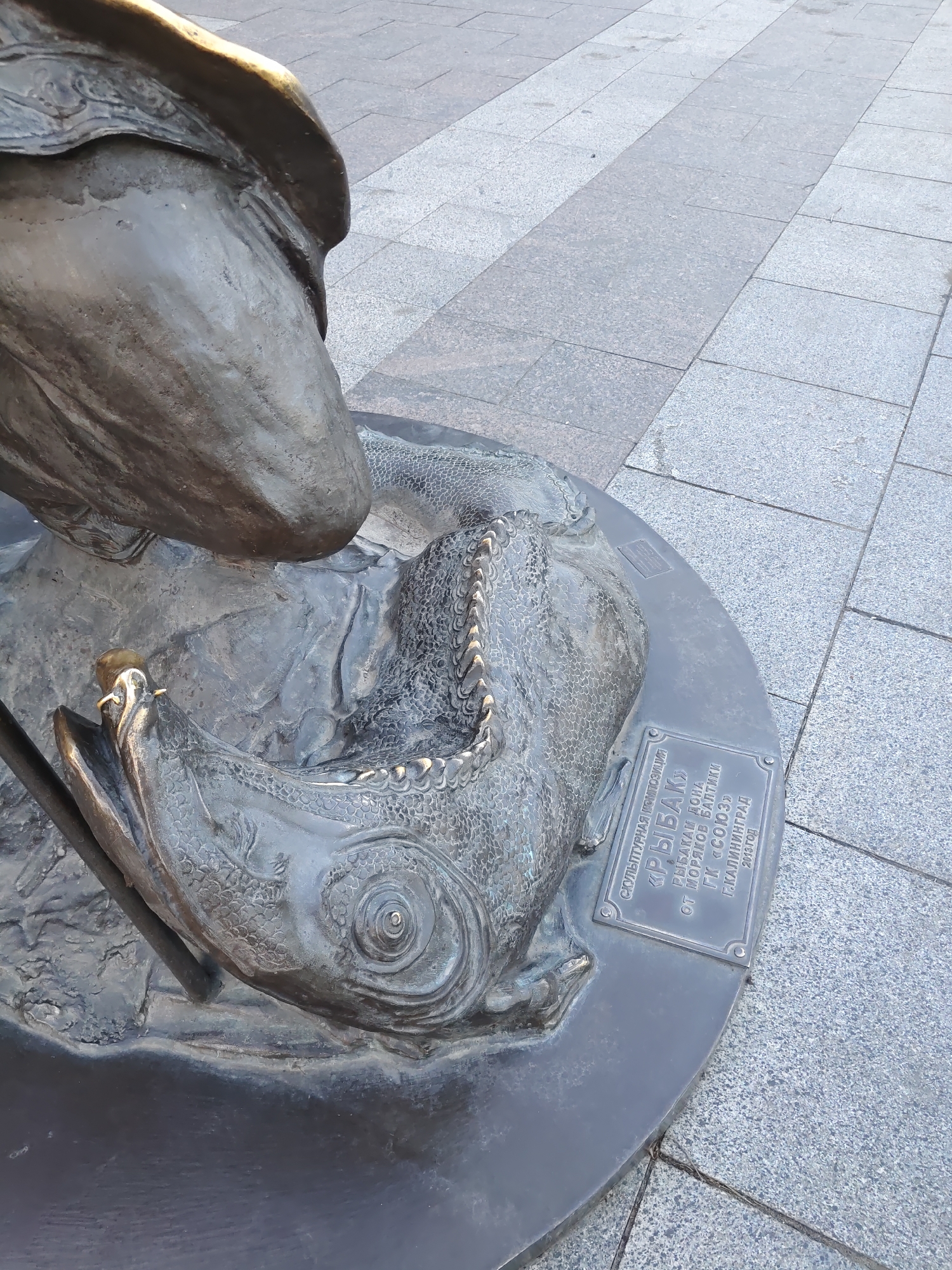 The image shows a close-up of a bronze sculpture located in Rostov-on-Don, Russia. The sculpture depicts a fisherman and a large fish. The fisherman's hand is visible, holding a fishing rod which is near the fish's mouth. The fish is intricately detailed with scales and fins. A plaque on the base of the sculpture reads "СКУЛЬПТУРНАЯ КОМПОЗИЦИЯ «РЫБАК» ОТ МОРЯКОВ БАЛТИКИ ГК «СОЮЗ» Г. КАЛИНИНГРАД 2013 ГОД". This translates to "Sculptural composition 'Fisherman' from Baltic sailors GK 'Soyuz' Kaliningrad 2013". The sculpture is situated on a paved ground made of large grey tiles. The lighting suggests it is daytime.
