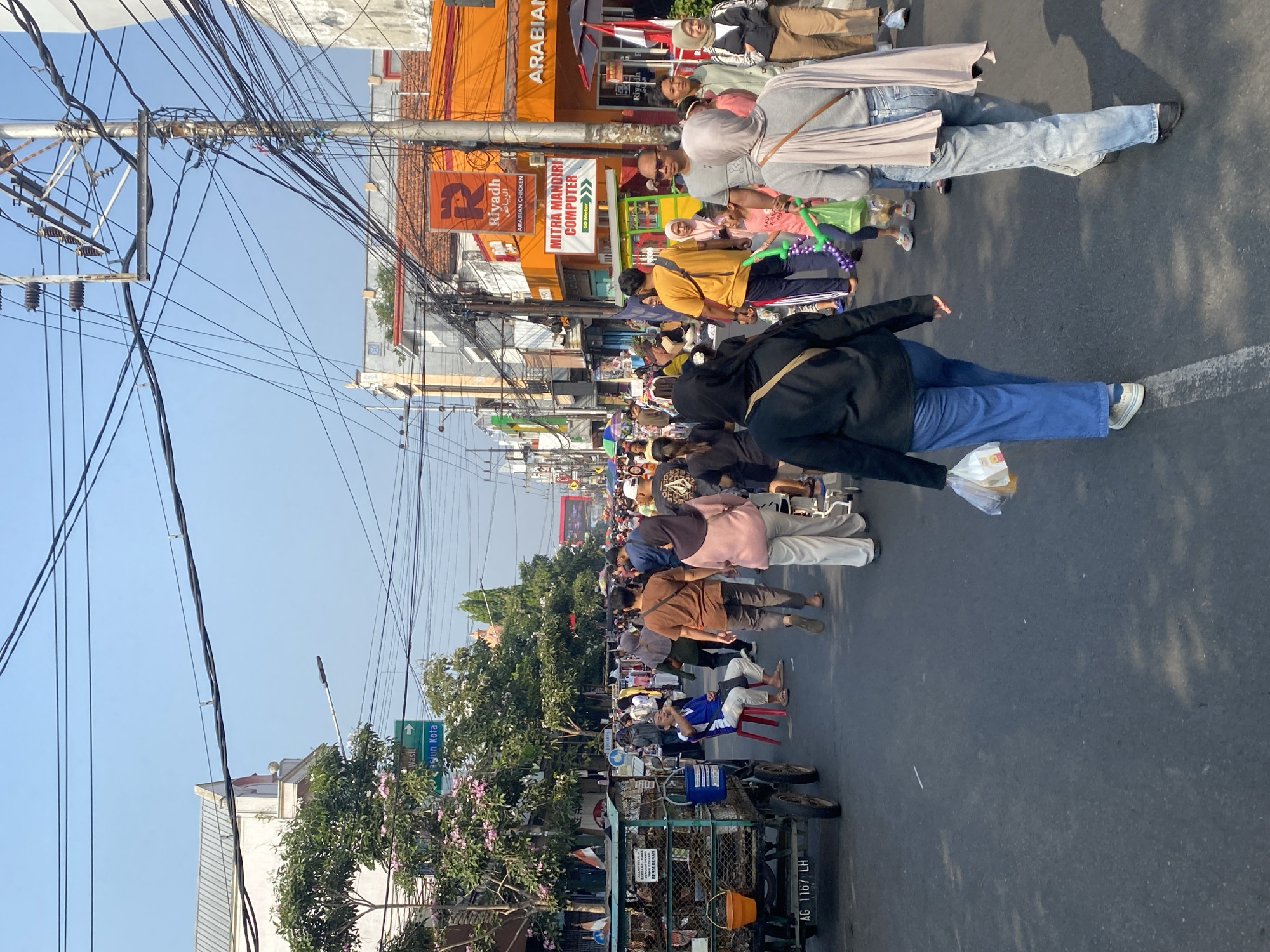 The image depicts a busy street scene in Kediri City, Indonesia. A crowd of people are walking on the asphalt road, with some individuals seated on chairs. Various stalls and shops line the street, featuring awnings and signage. One stall has a cart with baskets and buckets. A signpost indicates directions to "Wasil" and "Pasj'un Kota." Overhead, a network of electrical wires crisscrosses the sky, supported by utility poles. The buildings on either side of the street appear to be commercial establishments, with one prominently displaying an orange facade and the "Riyadh Arabian Chicken" logo. Another sign advertises "Mitra Mandiri Computer." The atmosphere appears to be that of a market or a bustling city street during daylight hours, with clear skies overhead.