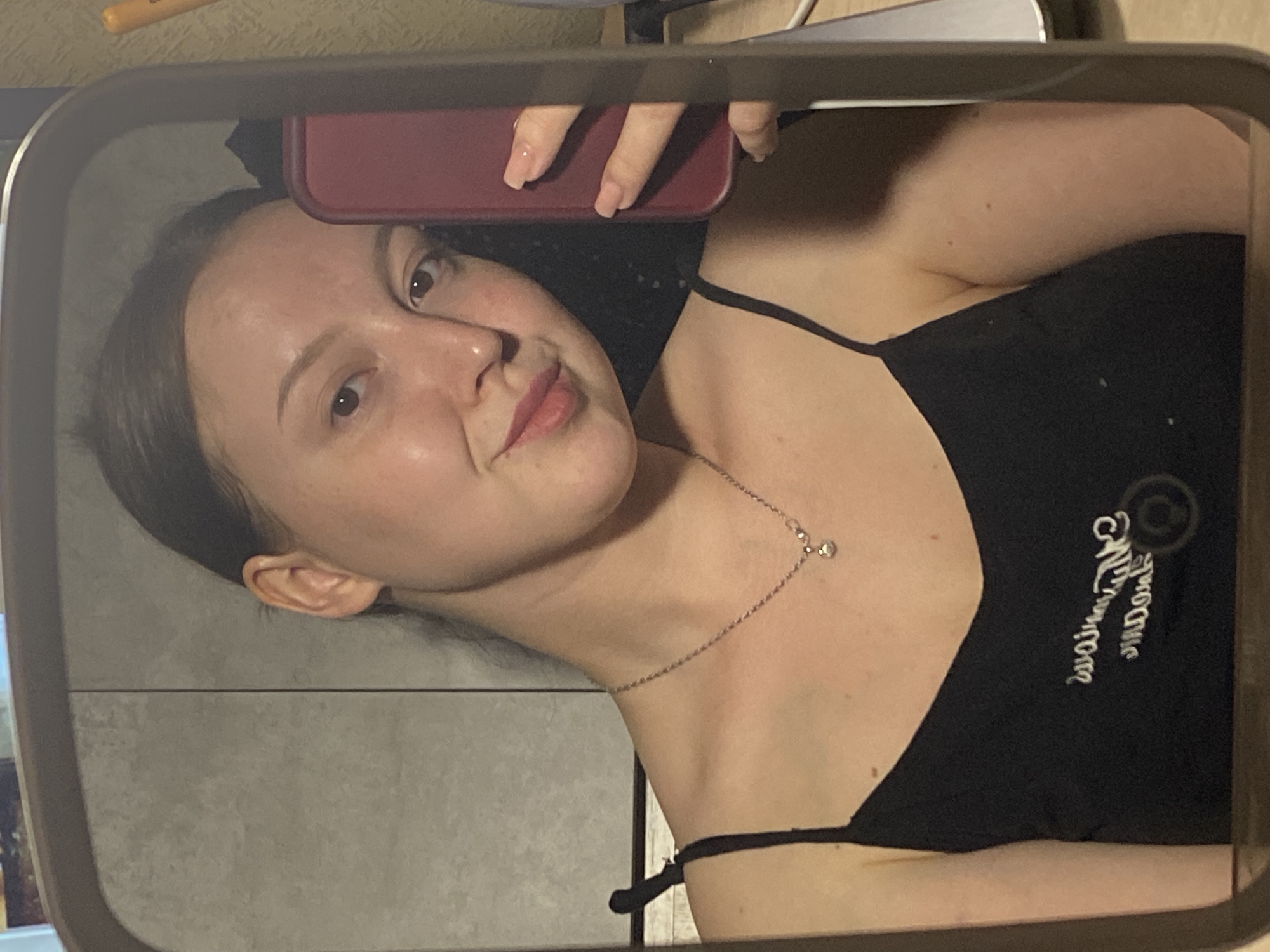 A woman is taking a selfie in a mirror. The woman has light skin, brown hair, and is wearing a black tank top and a silver necklace. She is holding a red smartphone. The background is a tiled wall. The location is likely Chaikovsky, Russia, based on analysis of social media patterns.