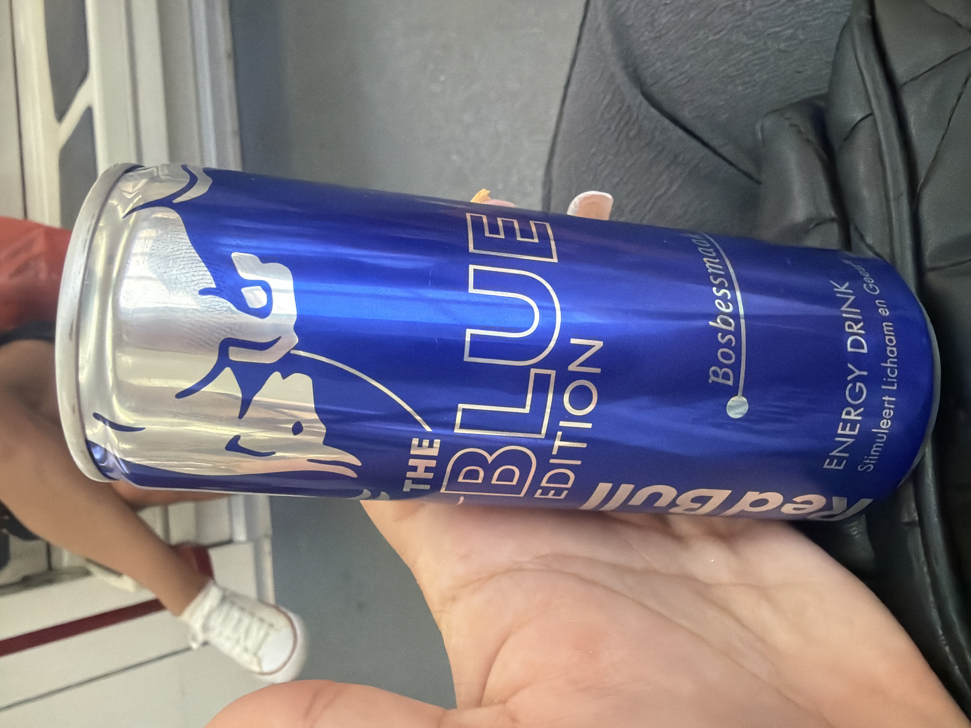 A person's hand is holding a can of Red Bull. The can is blue with silver lettering and features the Red Bull logo. The text on the can reads "THE BLUE EDITION" and "Bosbessmaak". Below this, it says "ENERGY DRINK" and "Stimuleert Lichaam en Gees". The person's leg with a white sneaker on is visible in the background, suggesting they are seated. The environment appears to be indoors, possibly on public transportation, with white and grey seating.