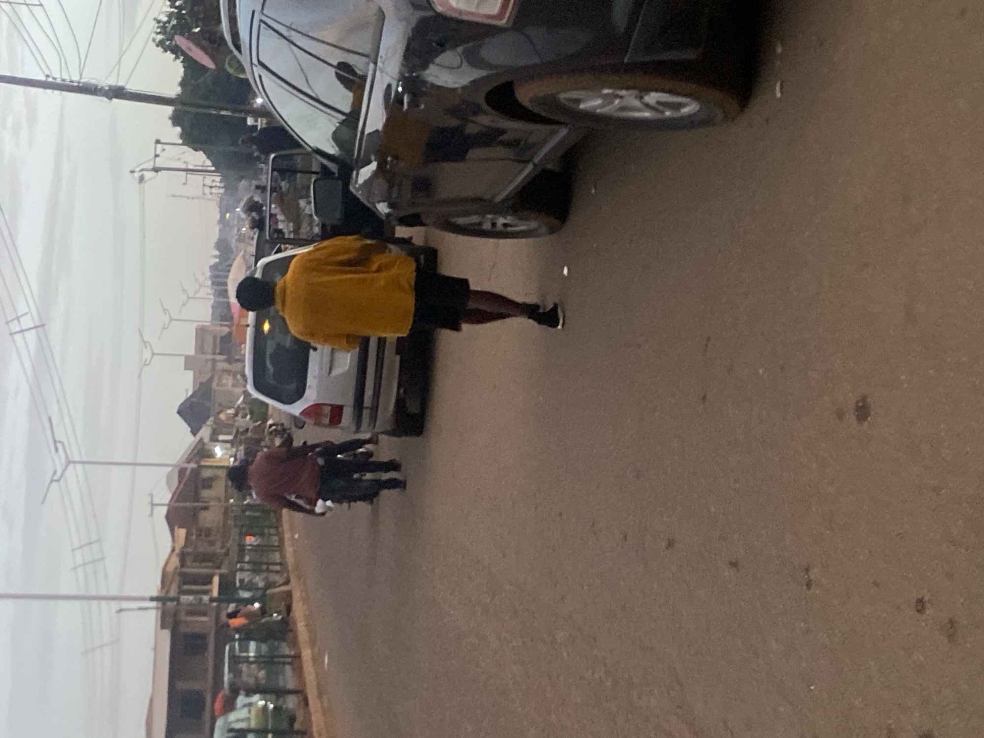 The image depicts an overcast street scene in Bukuru, Nigeria. The foreground features a reddish-brown unpaved or dirt road. On the right, a dark-colored SUV, possibly a Ford Edge, is partially visible, either parked or slowly moving, with its front right wheel and side facing the camera. A male individual in a bright yellow long-sleeved top and black shorts walks away from the camera on the right side of the road, wearing dark sneakers. In the center-left, a motorcycle is driven by a male in a reddish-brown shirt, carrying a passenger behind him, both moving away from the viewer. Behind the yellow-clad individual, a light-colored sedan or hatchback is visible, possibly parked.
In the midground on the left, a series of low-rise buildings, some with reddish roofs, are partially obscured by a dark-colored metal fence or barrier. Various other indistinct vehicles and people are visible along the roadside and in the distance. The background reveals numerous utility poles with overhead power lines. Distinctive streetlights with wide, flat heads, possibly indicative of modern or solar-powered designs, line the road stretching into the distance. The sky is uniformly overcast and light grey, diffusing the daylight. The distant horizon shows a continuation of buildings and a hazy urban landscape.