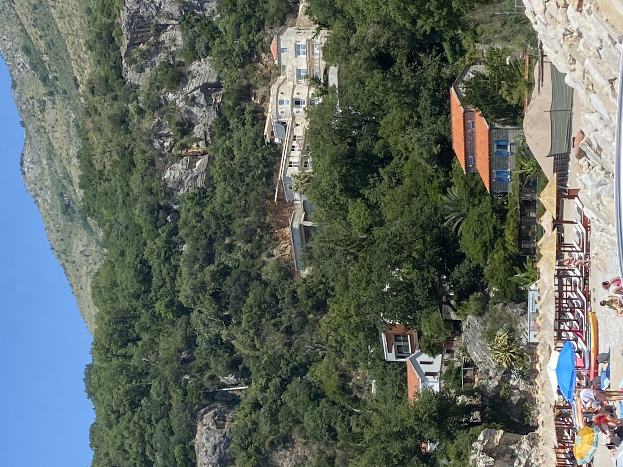 In Perazića Do, Montenegro, a sun-drenched beach scene unfolds with the verdant, mountainous terrain of the Adriatic coast serving as a dramatic backdrop. The foreground features a section of a rocky shoreline, with a sandy beach populated by numerous lounge chairs and thatched umbrellas, indicating a resort or public beach area. Several people are visible on the beach, some reclining on chairs, others standing or walking. A bright blue umbrella is open, providing shade. Kayaks and other beach equipment are also present.
Closer to the water's edge, a small group of individuals are gathered, with some appearing to be in swimwear. In the middle ground, nestled amidst dense green foliage, are several buildings. One prominent structure is a multi-story building with a light-colored facade and multiple balconies, appearing to be a hotel or residence. Further up the slope, another building with an orange tiled roof and blue shutters is visible, suggesting a smaller dwelling or guesthouse. The overall environment is characterized by lush vegetation, rocky outcrops, and clear blue skies, typical of the Mediterranean coastline. The buildings are integrated into the natural landscape, with their architecture complementing the steep, tree-covered hillsides.