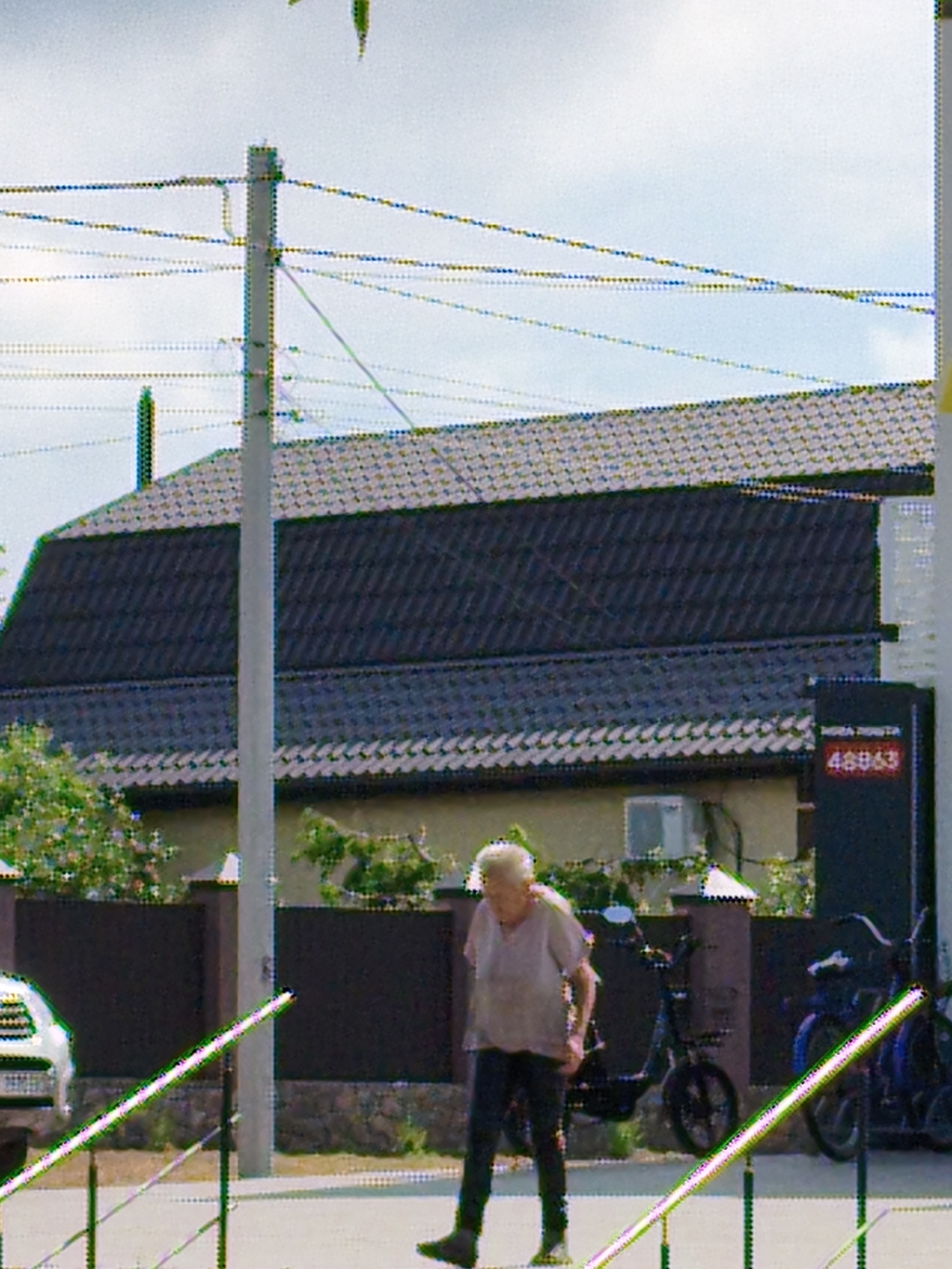 A woman wearing a light-colored short-sleeved shirt and dark pants stands on a sidewalk in front of a building with a dark tiled roof. She is facing left and appears to be looking towards something off-camera. To her right are two parked scooters. A white car is visible to her left. In the background, a sign with red text and numbers is attached to a dark pole. Overhead, electrical wires are strung between poles. The scene is outdoors with a bright sky.