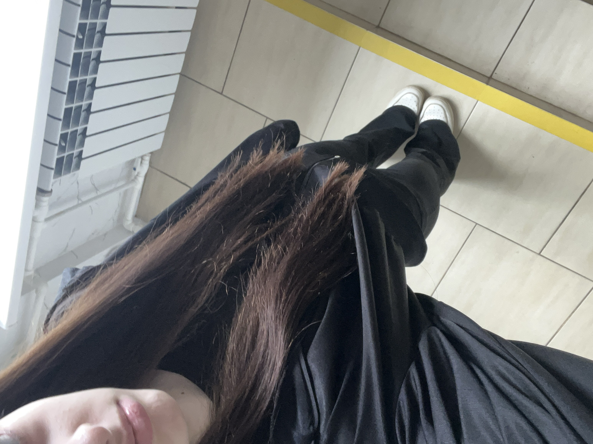 An aerial view of a person standing on a tiled floor with a yellow line marking a division. The person is wearing black pants and white sneakers. Part of the person's face and long brown hair are visible in the lower left corner. A white radiator is situated in the upper left corner. The location is Saint Petersburg, Russia.