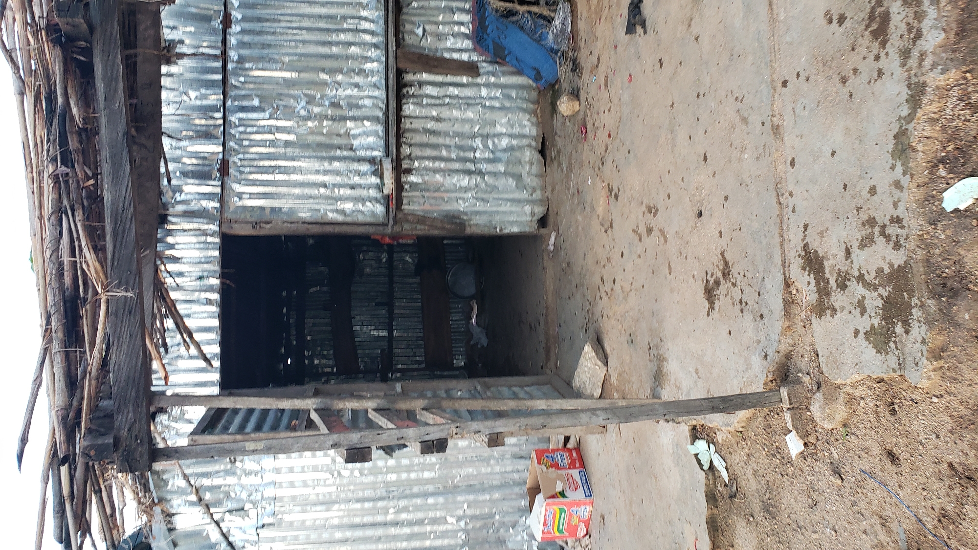 The image depicts the entrance to a makeshift structure in Jauro Yinu, Nigeria, constructed from corrugated metal and supported by wooden beams and branches. The roof appears to be made of layered branches and other natural materials. The entrance is open, revealing shelves inside the structure lined with what appear to be plastic containers or bags. The ground in front of the structure is concrete, stained and uneven, with scattered debris including a cardboard box with "Indomie" branding, a blue tarp, rocks, and what looks like discarded paper or plastic. The overall scene suggests a rural or impoverished setting.