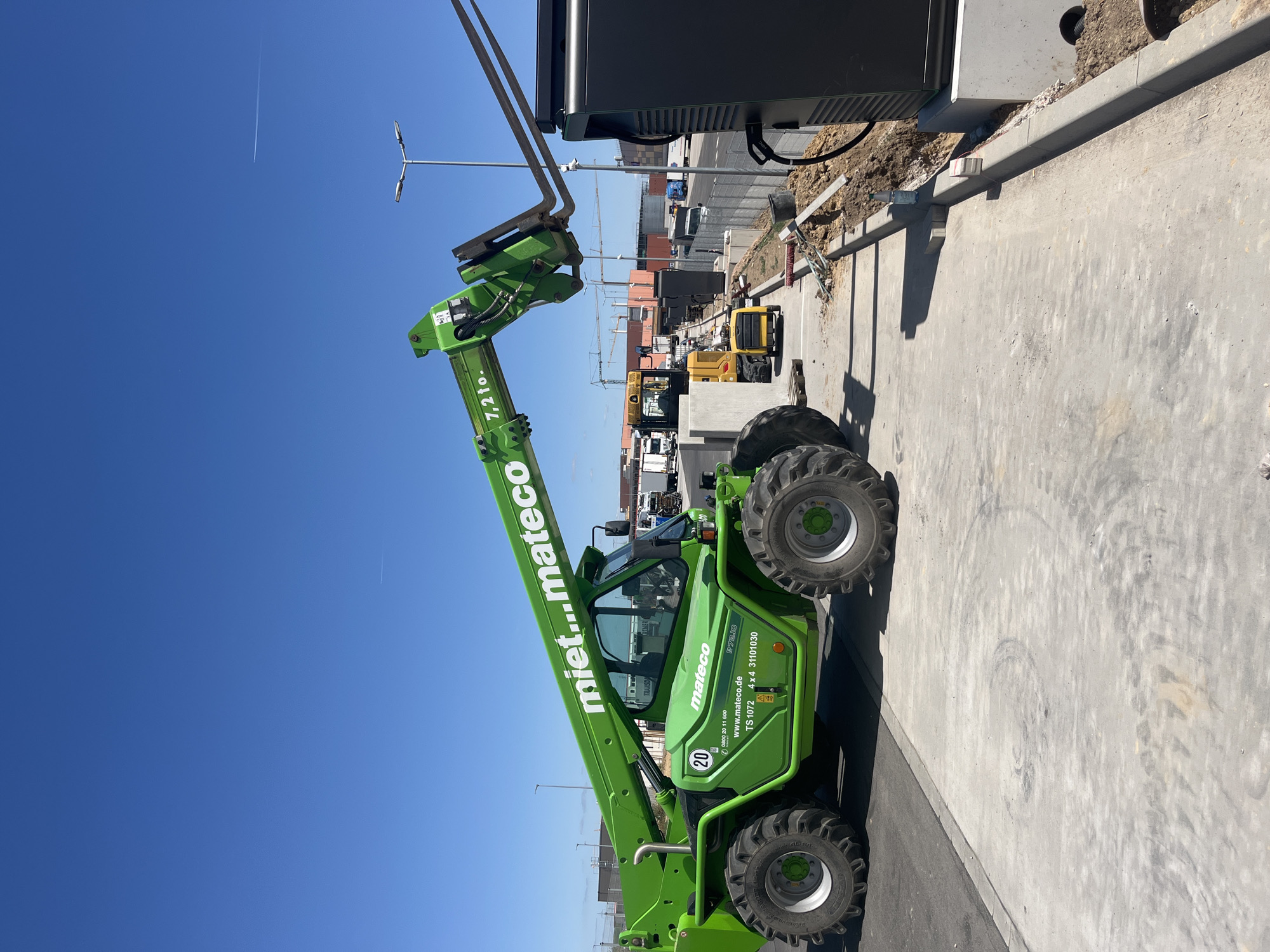 In Alzey, Germany, a bright green telehandler is parked on a paved surface. The telehandler's boom is extended upwards and slightly to the right, with its forks positioned near a tall, black electrical charging station. The charging station is mounted on a concrete base and has a thick black cable extending from it. The telehandler has "mi...mateco" written on its boom and "mateco" on its side, along with the model number "TS 1072 4x4 31101030" and the weight capacity "7,2 to.". Two large, knobby tires are visible on the left side of the telehandler. In the background, several trucks, construction equipment, and buildings are visible under a clear blue sky with a faint contrail. The ground is a mix of concrete and dirt, with some construction debris scattered around.