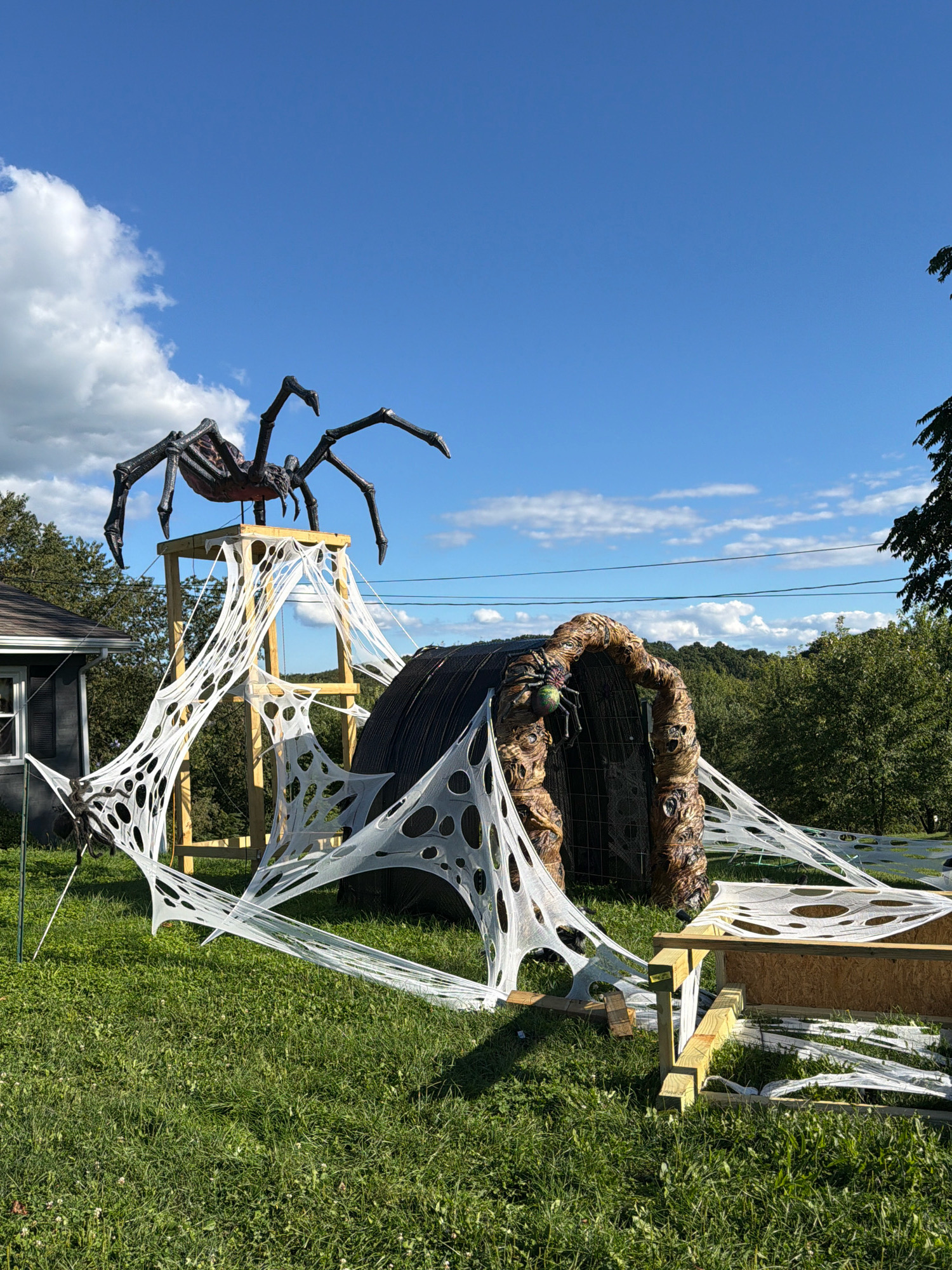 In North Sewickley Township, United States of America, a Halloween-themed outdoor decoration display is set up on a grassy lawn. A large, dark-colored spider decoration is perched atop a wooden platform, which is supported by wooden beams. White, tattered fabric resembling spiderwebs stretches from the platform and across the scene, connecting to various structures. To the right of the platform, a large, arched tunnel-like structure with a textured, earthy exterior is present. A small, colorful spider decoration is visible on the left side of this tunnel. A portion of a dark-colored building with a window is visible on the far left of the image. The background features a line of trees and a bright blue sky with scattered white clouds.