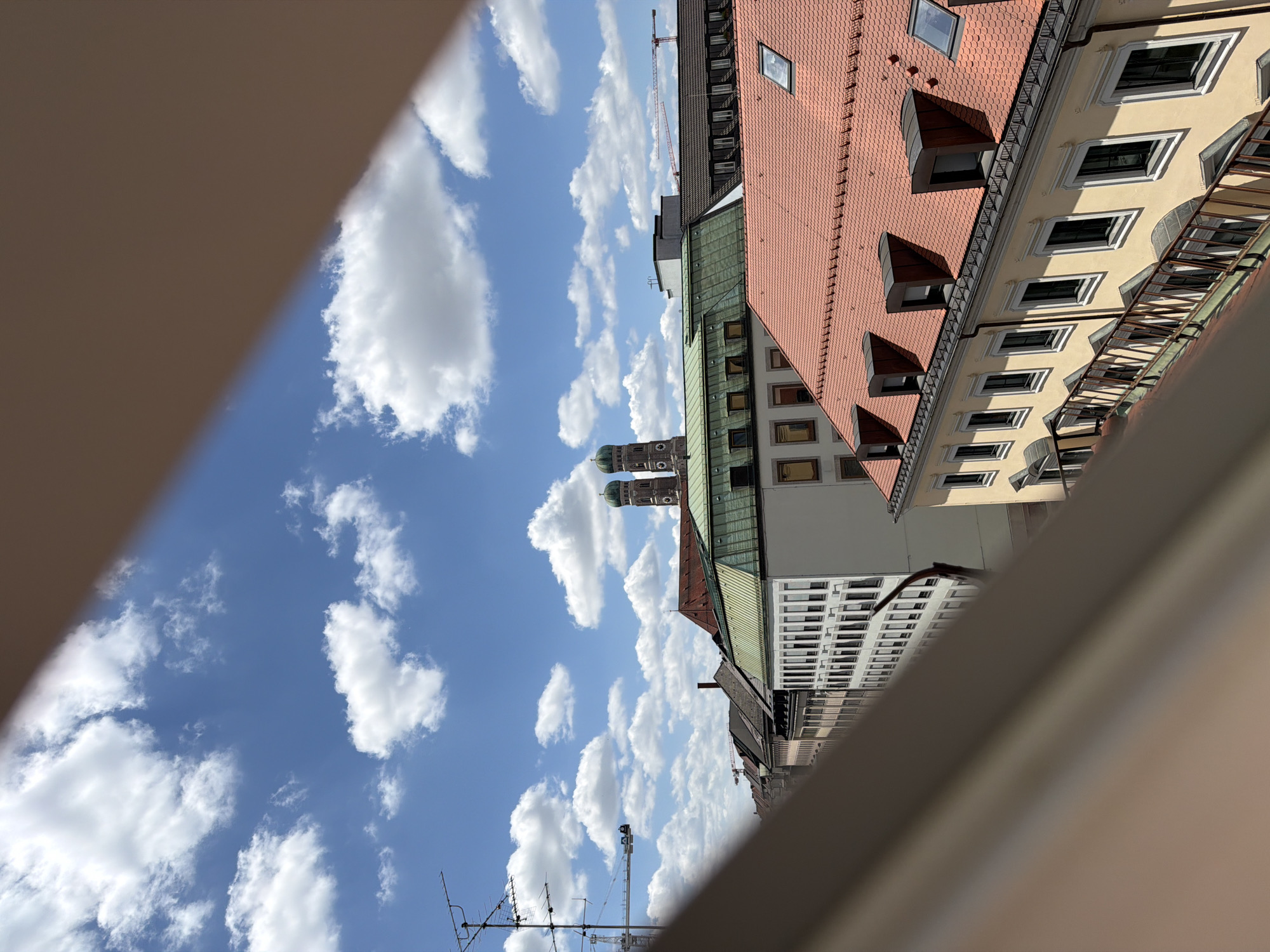 The image shows a view of the Frauenkirche (Church of Our Lady) in Munich, Germany, from a rooftop vantage point. The iconic twin green domes of the church are visible above a line of buildings with red tile roofs and lighter colored facades. The sky is blue with scattered white cumulus clouds. In the foreground, the edge of a structure, likely a balcony or railing, is visible, casting a shadow and partially obscuring the view. Other buildings with various rooflines and window arrangements are visible in the midground, and a construction crane is present in the distance. The overall scene depicts a sunny day in a European city.
