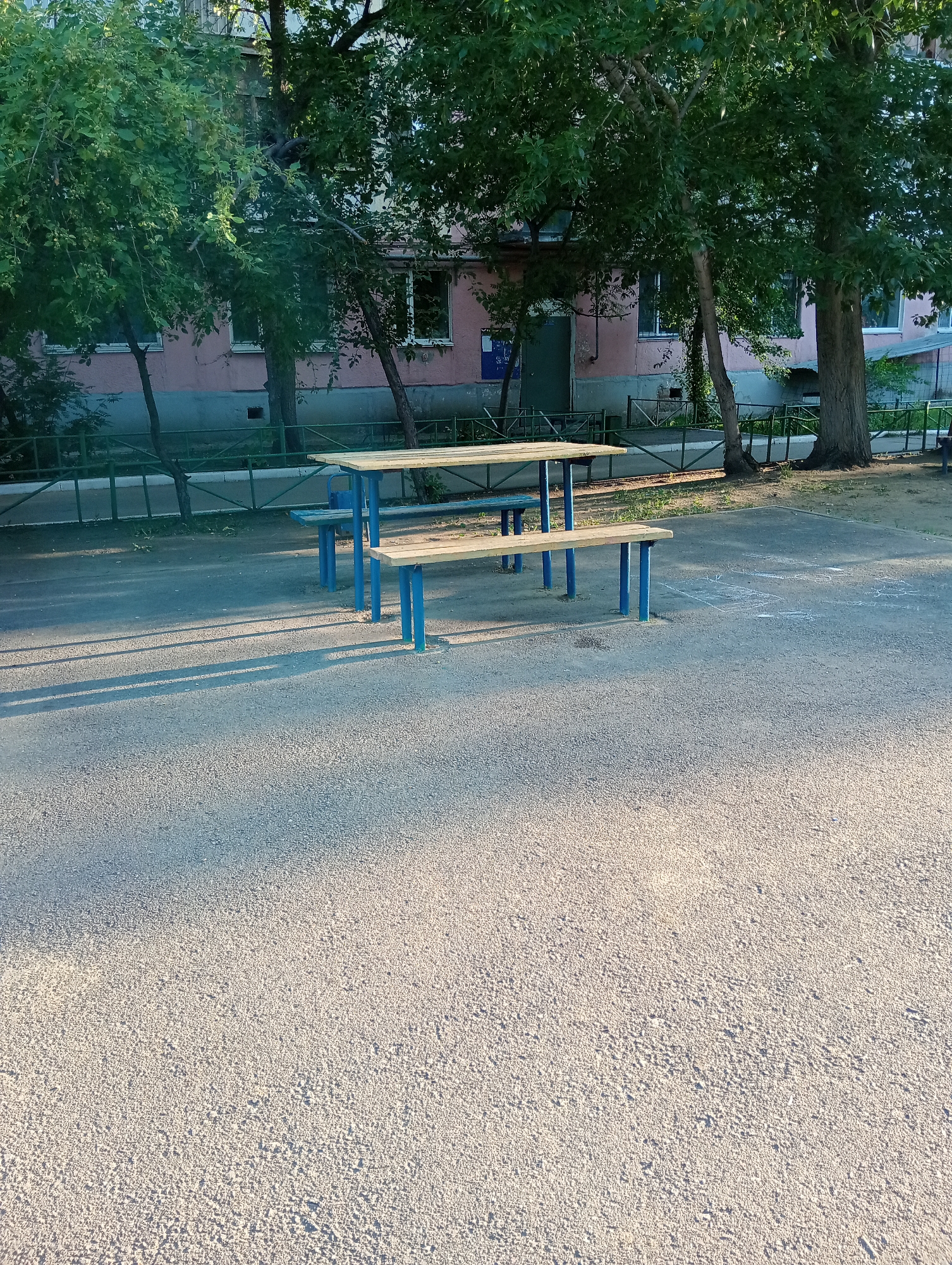 The image depicts an outdoor scene, likely a schoolyard or park, in an urban setting. In the foreground, a paved surface with a rough texture is visible, casting long shadows from an unseen light source, suggesting late afternoon or early morning. Two wooden picnic tables with blue metal legs are positioned on the paved area. Behind the tables, a row of trees with dense green foliage partially obscures a light pink building with multiple windows. A green metal fence runs along the edge of the property, separating it from a grassy area. The building appears to be residential or institutional. The overall impression is one of a quiet, somewhat dated public space.