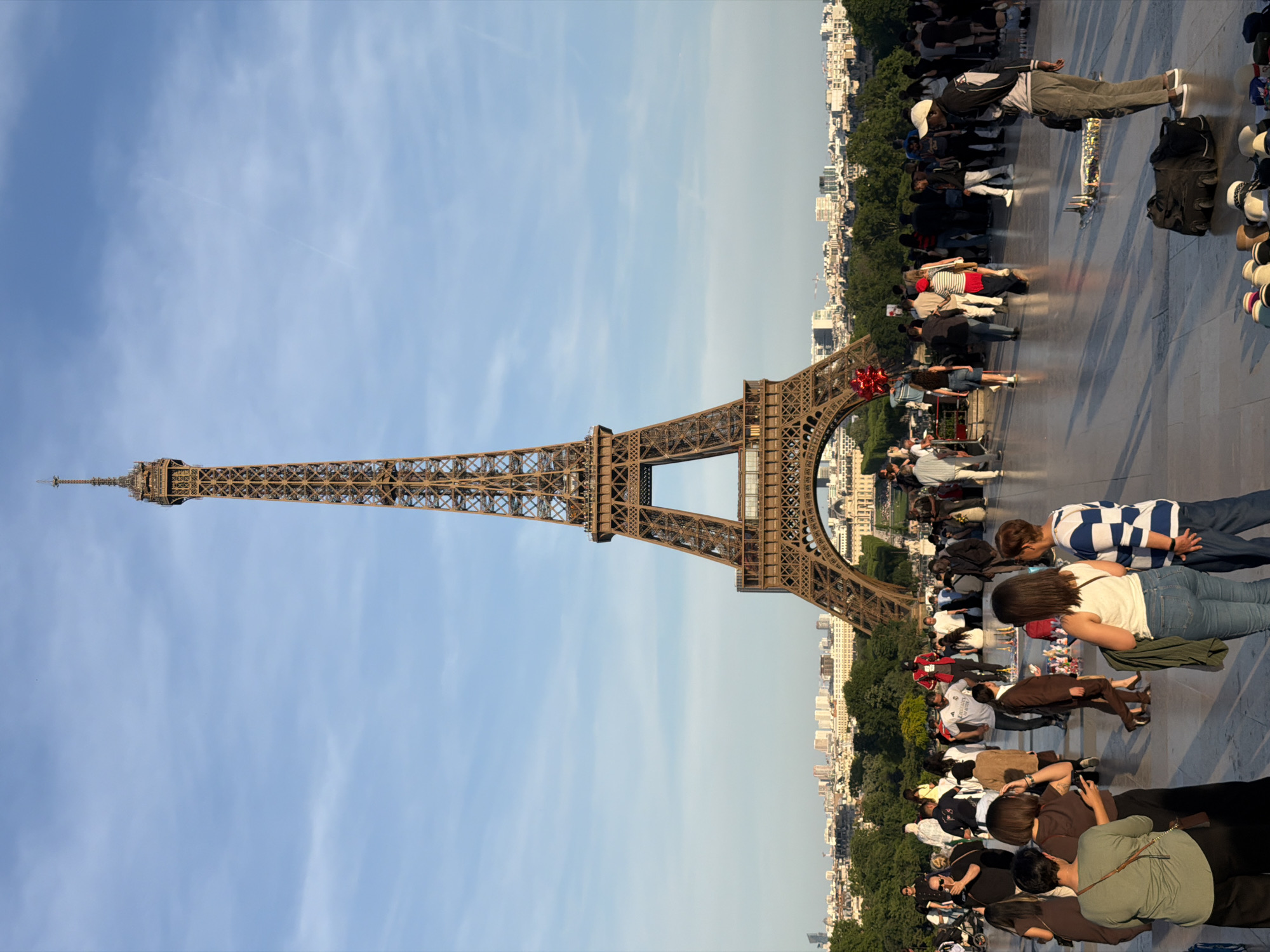 The image shows the Eiffel Tower in Paris, France, standing prominently against a blue sky. Many people are present, scattered around the base of the tower, likely tourists taking pictures. The surrounding city skyline is visible behind the tower, mixed with trees and buildings. The ground is paved.