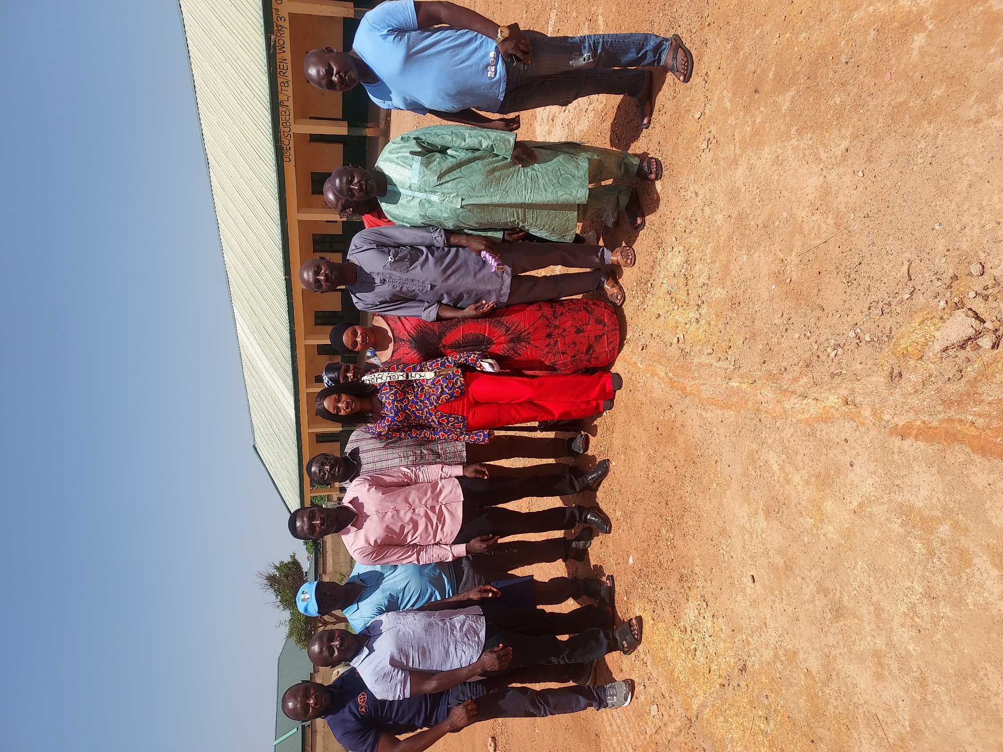 A group of approximately twelve individuals is standing in a line outdoors, presumably for a photograph. They are standing on what appears to be a dirt or sandy ground. The group includes both men and women, dressed in a variety of clothing styles, ranging from casual shirts and trousers to more traditional African attire. Behind the group is a building with a green roof and a wall that has writing on it, including "UBEC/SUBEB/PL/TB/REN WORKS 3rd". The sky is clear and blue. The image was likely taken during daylight hours.
Although the prompt mentioned the city of Jos, Nigeria, no detail in the image itself definitively confirms this location. Therefore, the description refrains from stating that the image is specifically in Jos, Nigeria.