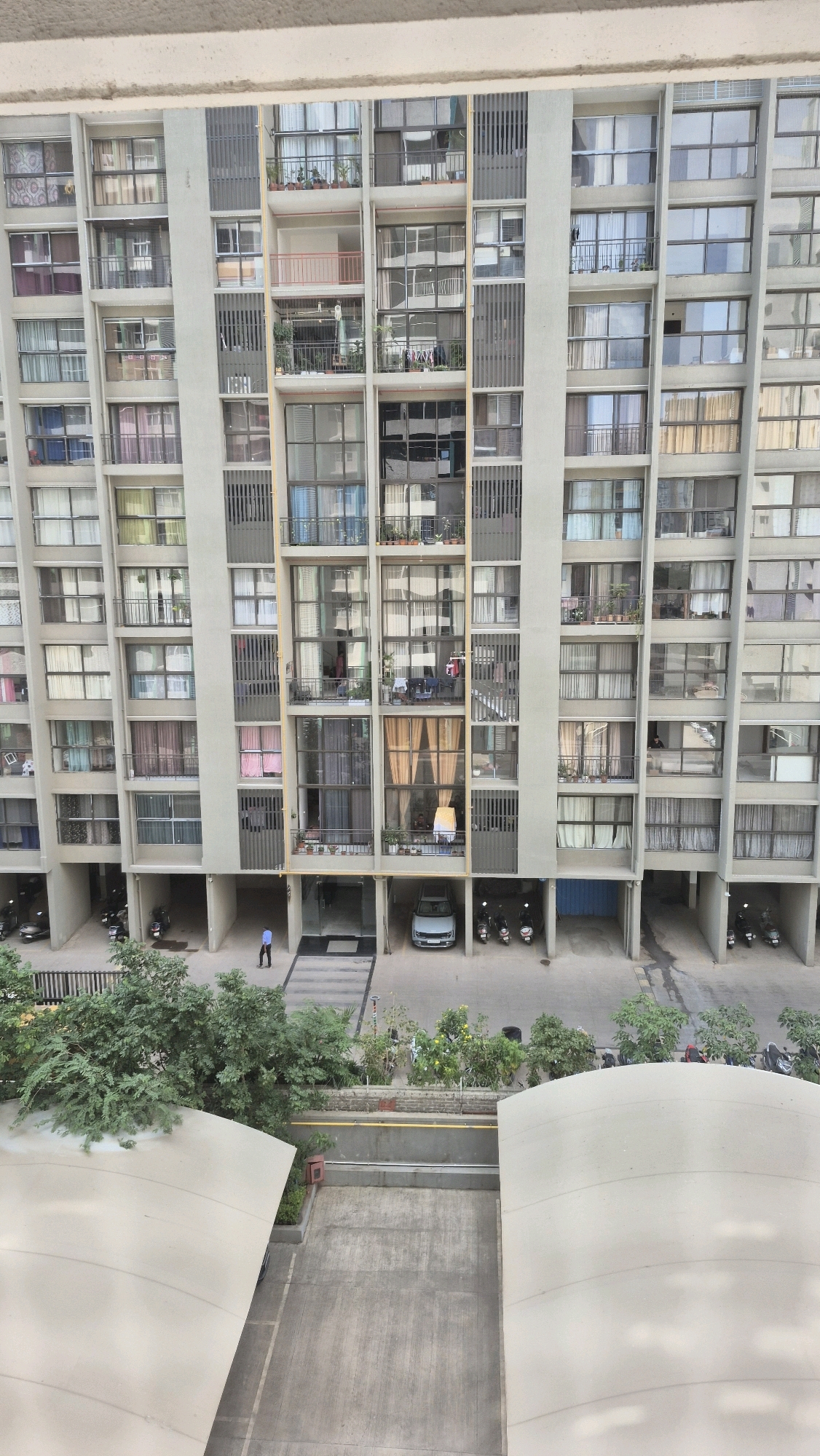 The image shows a tall, modern apartment building in Pune, India. The building has many floors, each with numerous windows and balconies. Some balconies have plants, and laundry is visible hanging on some. On the ground level, there is a parking area with a few cars and scooters. A single person in a blue shirt and dark pants is walking in front of the building entrance. Trees and greenery surround the base of the building. The overall scene depicts a typical urban residential area in Pune.