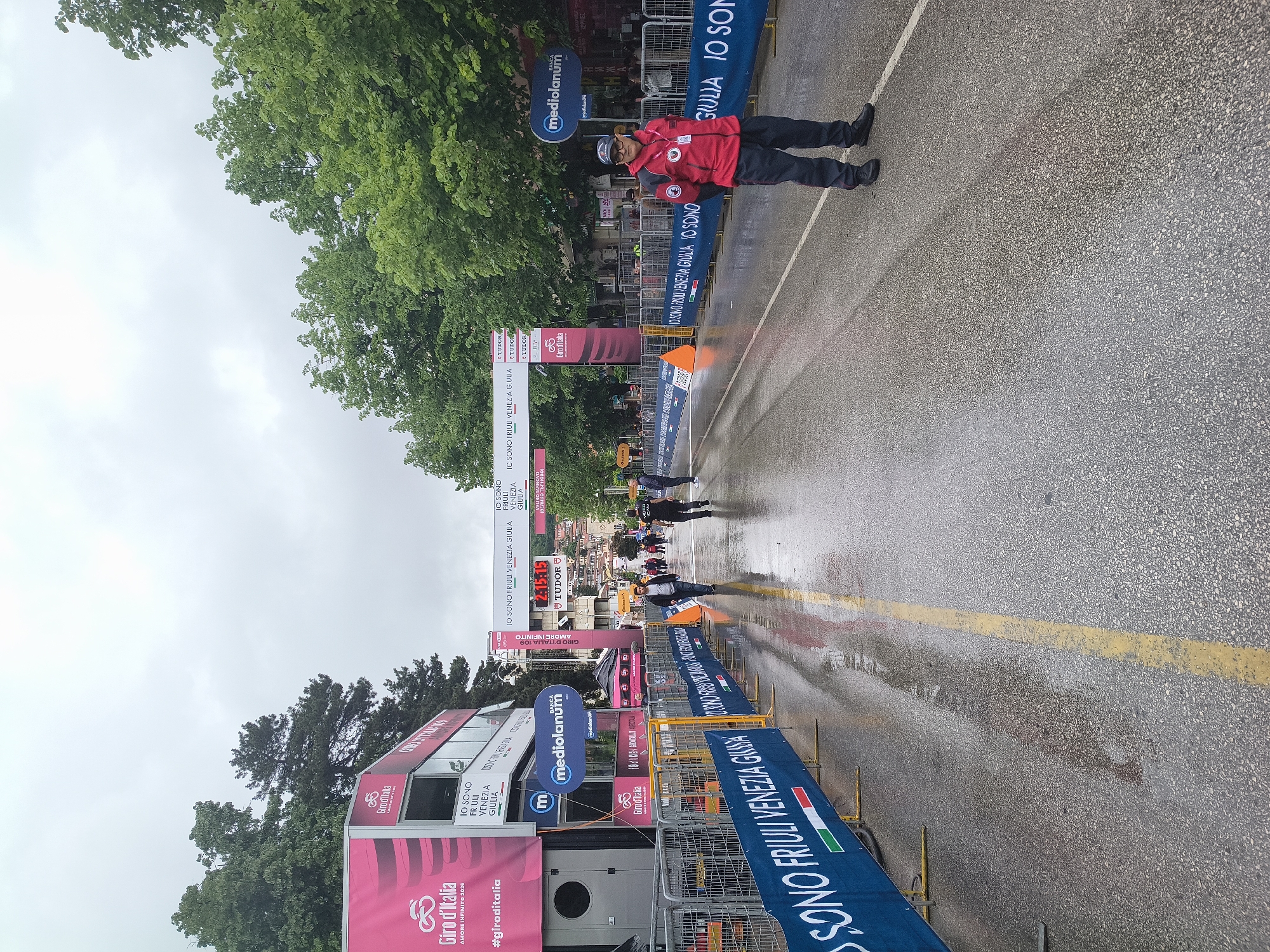 The image shows a finish line setting of a Giro d'Italia cycling event in what appears to be a wet urban environment. There is a man in a red jacket standing near the barrier on the right side of the frame. Multiple branded banners and structures are visible. The ground is wet, suggesting it has been raining. The city of Veliko Tarnovo, Bulgaria is not visible in the image.
