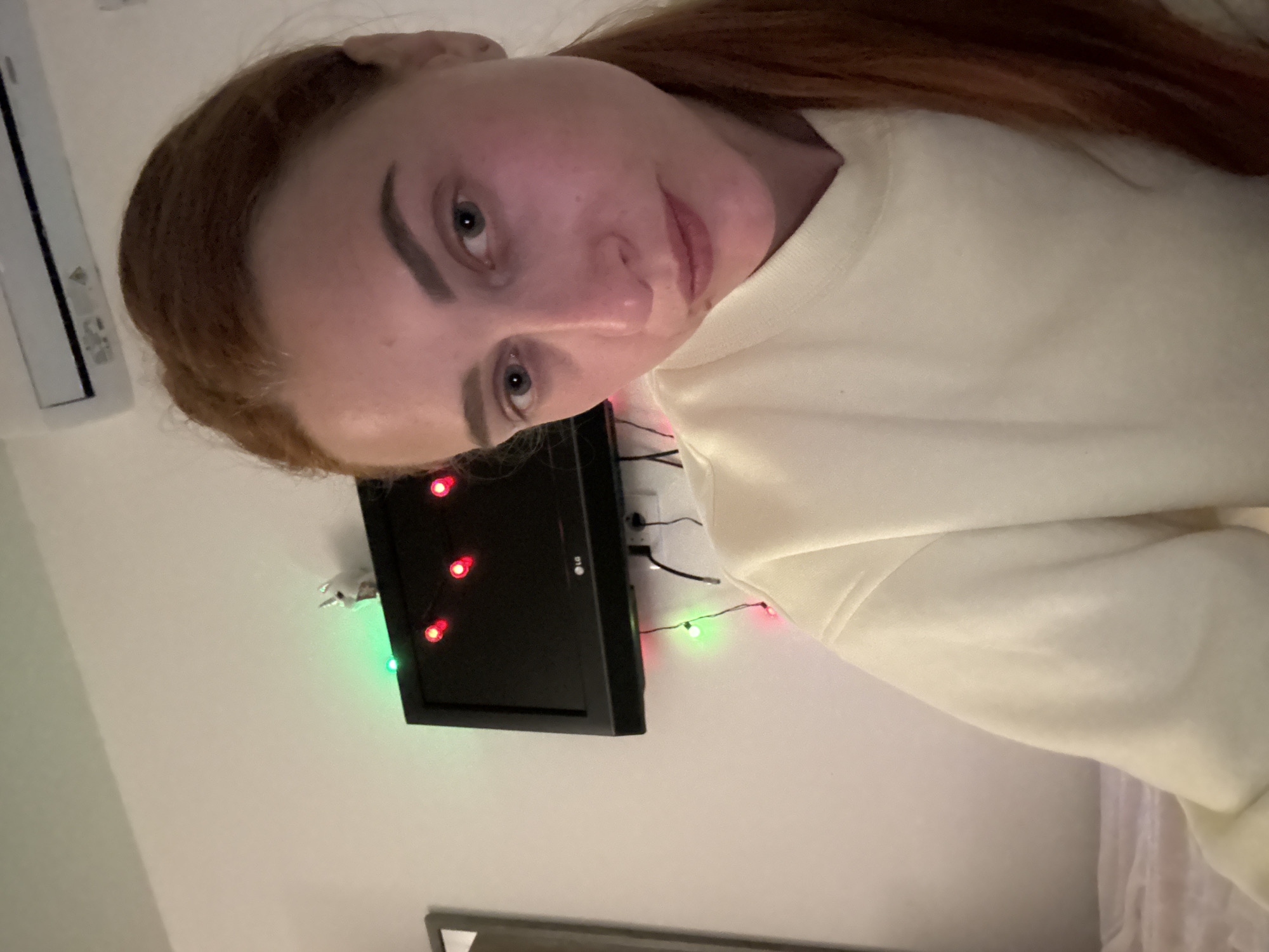 A woman with light skin and reddish-brown hair pulled back in a ponytail is shown in a close-up, angled self-portrait. She is wearing a cream-colored sweater. Behind her, mounted on a white wall, is a black LG television with red and green string lights attached to its frame. To the left of the TV is what appears to be an air conditioning unit, also mounted on the wall. Below the TV, a portion of a framed picture is visible. The setting appears to be indoors, possibly a residential room. The city of Budva, Montenegro cannot be confirmed or denied based on the provided image alone.