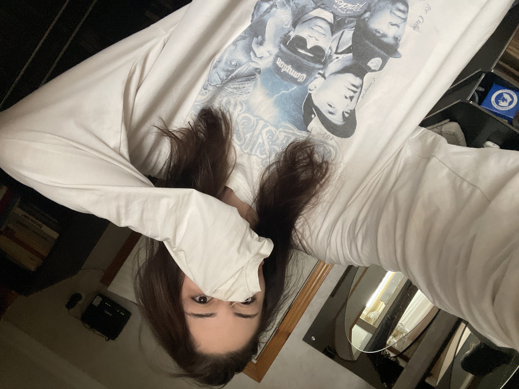 A young woman with long, dark hair is taking a selfie in what appears to be her bedroom. She is wearing a white, long-sleeved t-shirt with a graphic print of several men, some wearing hats. Her face is partially obscured by the sleeve of her shirt as she covers her mouth and nose with it. Only her eyes and eyebrows are clearly visible, and she is looking directly at the camera. The background shows a dimly lit room with a mirror on the left side, reflecting part of the room. There is also a shelf with books and a router visible in the background. The overall lighting suggests it is nighttime or the room is dark. There are no discernible elements that would place the location in Moscow, Russia.