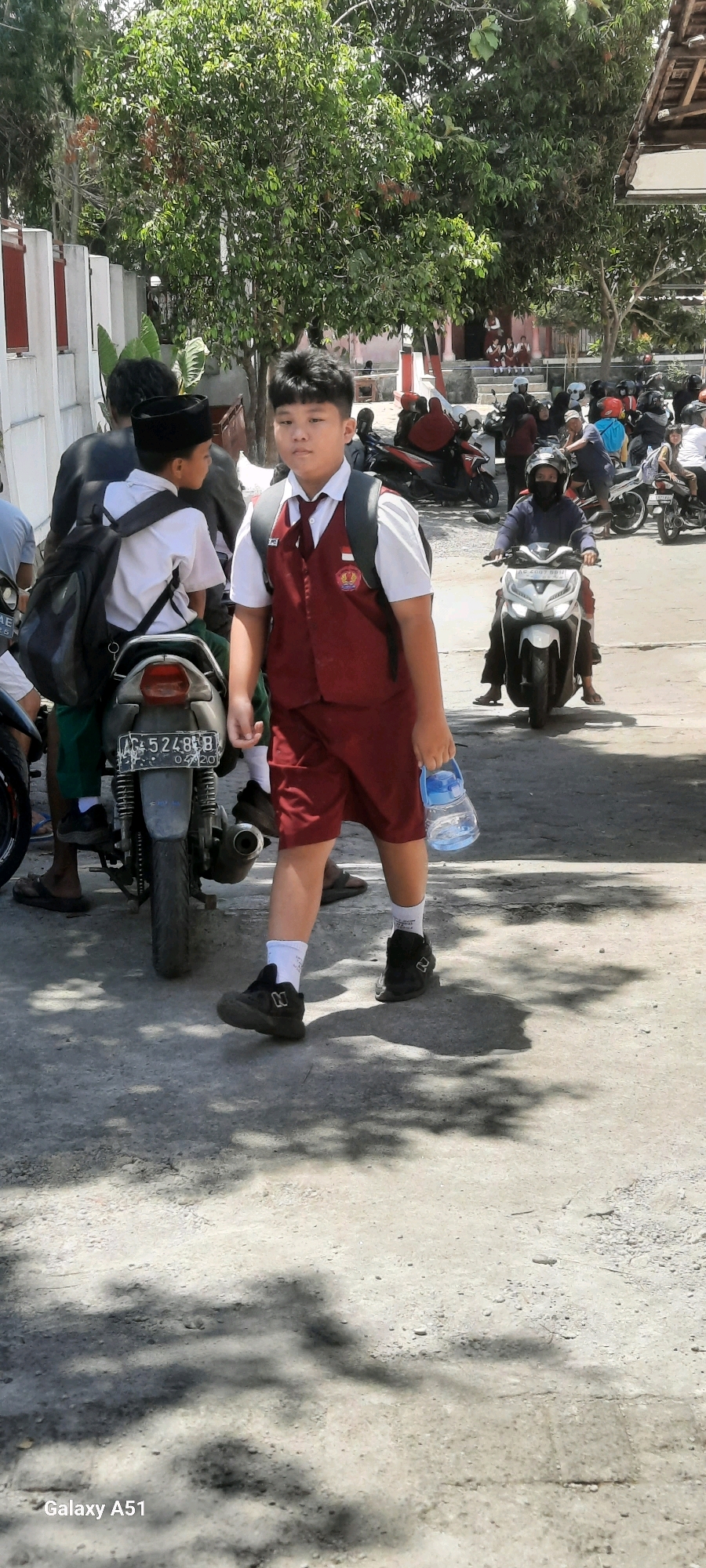 In Tangkil, Indonesia, on a bright, sunny day, a young male student, dressed in a white shirt, maroon vest, maroon shorts, white socks, and black shoes, walks forward, holding a blue water bottle. He has short, dark hair and is carrying a dark backpack. Behind him, another student, wearing a black skullcap, white shirt, and green pants, sits on a motorbike. Several other motorbikes and people are visible in the background, some wearing helmets, suggesting a busy street or parking area. The ground is paved, and dappled shadows from trees fall across it, indicating an outdoor setting. The overall scene depicts a moment of everyday life with students preparing to depart or arrive, likely at a school in the area.