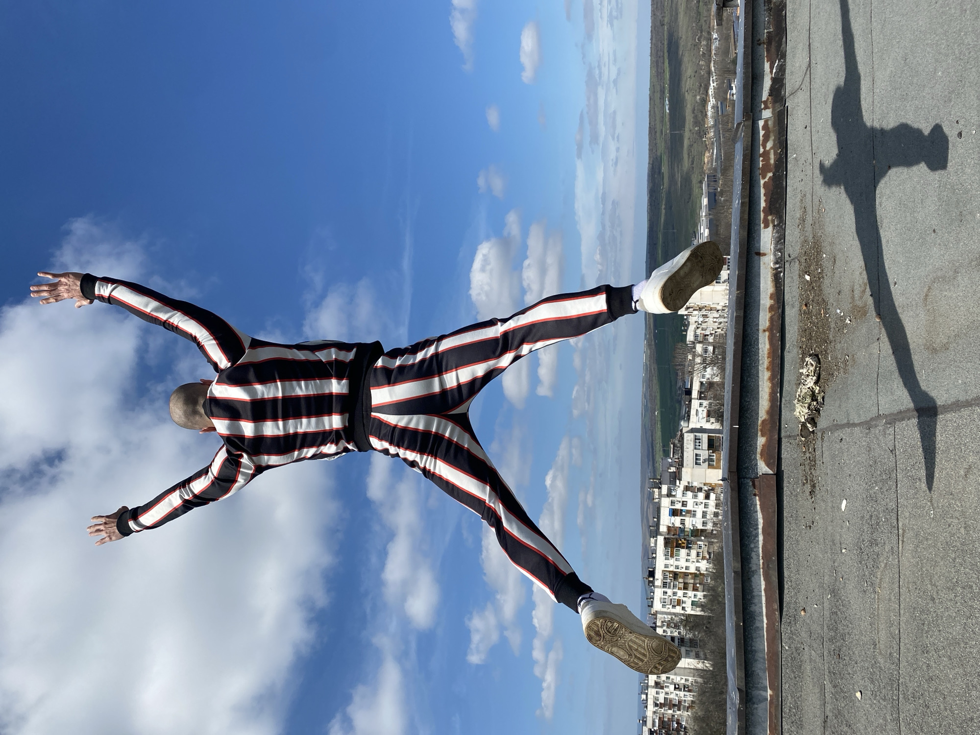 A photograph shows a person lying horizontally on the edge of a rooftop, with their arms and legs outstretched. The person is wearing a striped jumpsuit with black, white, and red lines. They are wearing white sneakers. The person is bald. The background features a bright blue sky with scattered white clouds. Below the rooftop, there is a view of a city. The shadow of the person is visible on the rooftop. The image may have been taken in Pleven, Bulgaria, given the presence of typical buildings in the background. The photo is taken at an angle, giving the impression of height and depth.
