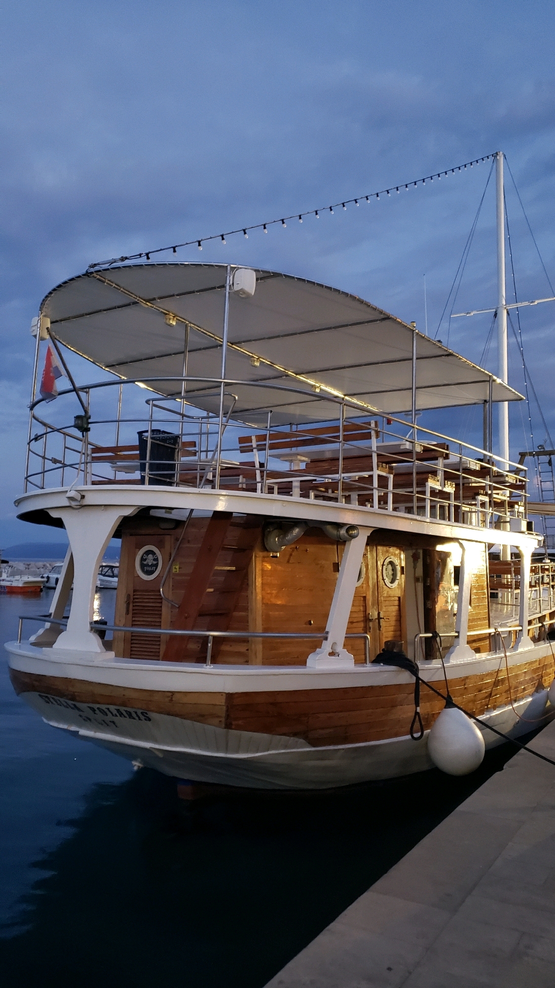 The image shows a wooden boat named "STELLA POLARIS" docked in a harbor, likely in Općina Tučepi, Croatia. The boat has multiple levels with an upper deck covered by a canvas awning, supported by metal poles. The deck features wooden benches and tables, suggesting it's used for passengers. String lights are visible along the edge of the awning. The boat has a traditional wooden hull with white trim and several porthole windows. A white fender is attached to the side of the boat by a rope, and another rope is tied to the dock. The water is dark and calm, reflecting the boat and the twilight sky. In the background, other boats and a hazy shoreline are faintly visible. The sky is cloudy and shows the transition from day to evening.