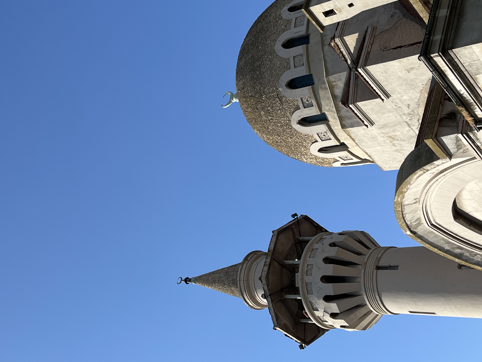 The image shows a close-up view of the Grand Mosque of Constanța, also known as the Carol I Mosque, located in Constanța, Romania. The photograph captures the upper portions of the mosque, focusing on the minaret and the main dome against a clear blue sky.
The minaret, on the left, is a tall, cylindrical tower with a conical top. It is made of light-colored stone and features intricate architectural details, including arched windows and decorative elements around its upper section. A crescent moon symbol tops the minaret.
To the right, the main dome of the mosque is visible. It is a large, rounded structure with a textured surface, possibly made of small stones or tiles. The dome is adorned with a series of arched windows and decorative motifs. A prominent crescent moon symbol is also situated on top of the dome.
The main building of the mosque, beneath the dome, is constructed from similar light-colored stone. It exhibits a combination of architectural styles, with arched windows and a layered roofline. The overall impression is of a majestic religious edifice bathed in sunlight. The scene is serene, with no people or any other discernible objects or actions in the frame, emphasizing the architectural grandeur of the mosque.