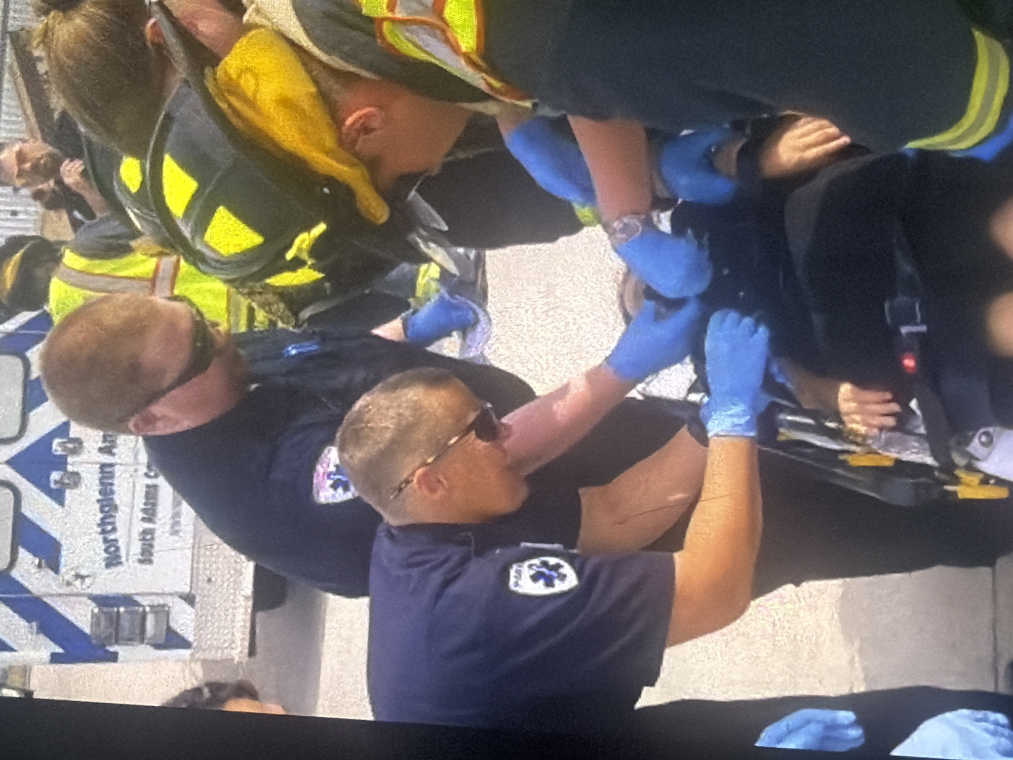 The image shows an emergency medical response scene. Several individuals in uniform, appearing to be paramedics and possibly firefighters, are attending to a person lying on a stretcher. The person on the stretcher is being attended to by multiple individuals wearing blue gloves. An ambulance with the markings "Northglenn An" and "South Adams C" is visible in the background, suggesting this is a professional medical intervention. The environment appears to be outdoors, possibly on a street or parking lot, with a textured ground surface. The focus is on the coordinated efforts of the responders to care for the individual on the stretcher. There is no information in the image to indicate this scene is in Bukuru, Nigeria.
