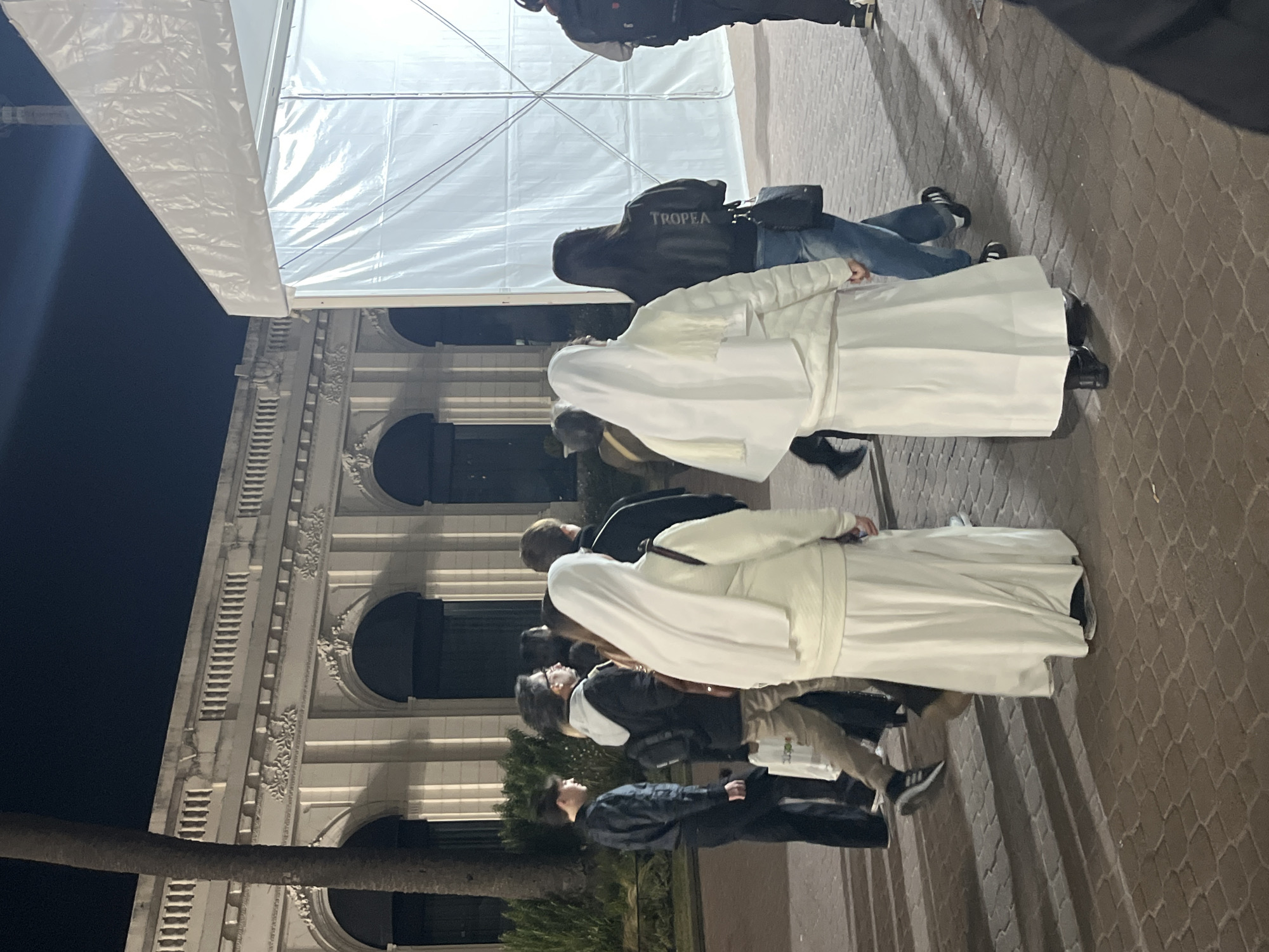 People are walking on a brick sidewalk beside a building with ornate decorations. Two people are wearing white garments, possibly religious robes. The background includes a tent-like structure, and a palm tree is partially visible. A person is wearing a jacket with the word "TROPEA" on it. It is nighttime. It is not possible to pinpoint the specific location in Buenos Aires without more information.