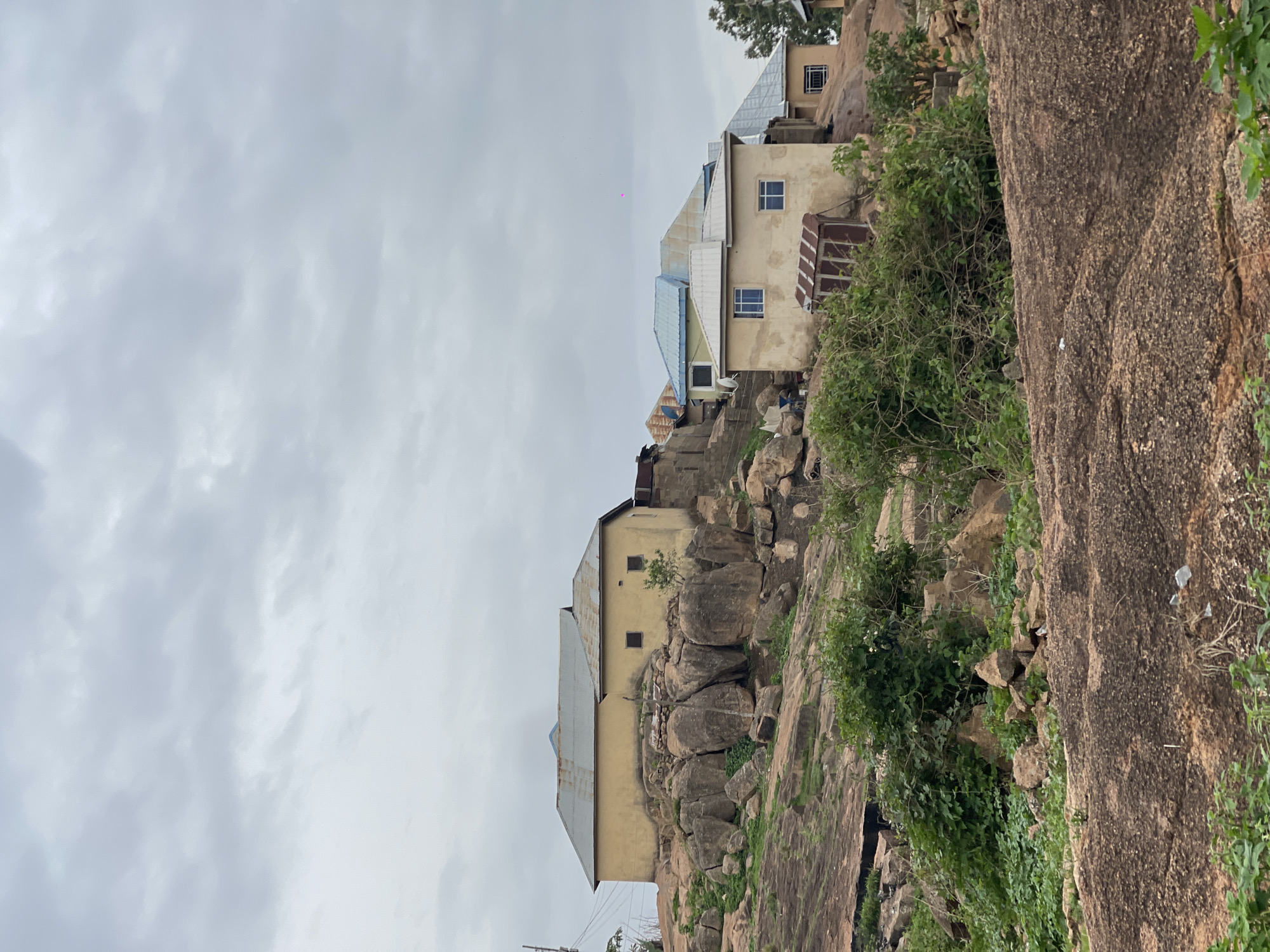 The image depicts a residential area in Jos, Nigeria, characterized by buildings constructed on and among large rock formations. Several structures are visible, ranging in size and design, with corrugated metal roofs and light-colored walls. The buildings are nestled into the natural landscape, integrating the rocks into their foundations and surroundings. Vegetation is abundant, with green foliage clinging to the rocks and interspersed among the buildings. The sky is overcast, casting a soft, diffused light over the scene. Power lines are visible.