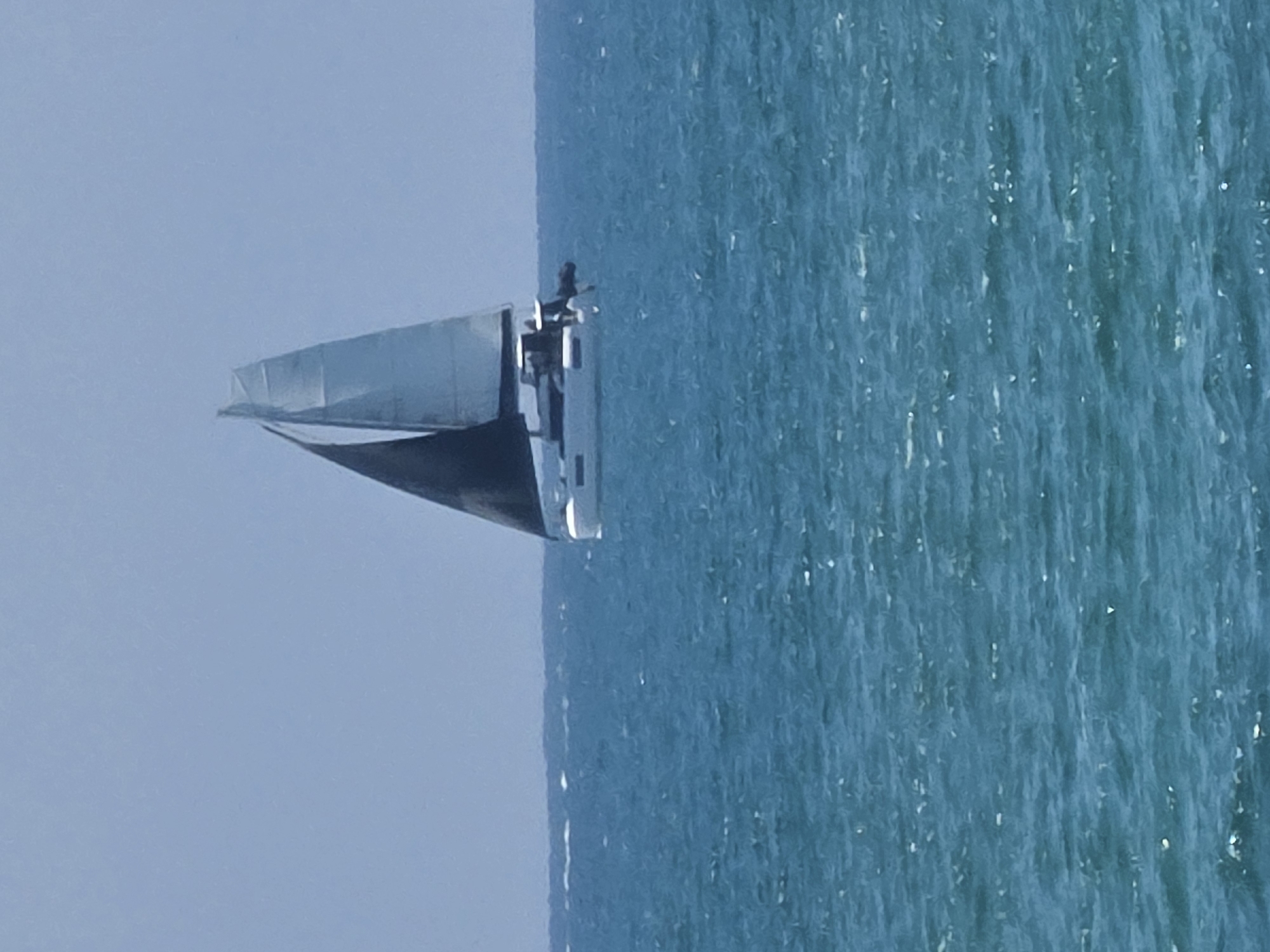 A sailboat is captured cruising on the calm, turquoise-blue waters under a clear, light blue sky, likely off the coast of Tel-Aviv, Israel. The boat, oriented from left to right across the frame, features a dark hull and a lighter-colored superstructure, including a cabin and deck. A prominent, light-toned mast or a furled wing-like sail structure is visible towards the bow. A small, dark figure, presumably a person, stands on the boat's deck near the stern, overlooking the water.
The sea exhibits gentle ripples and numerous sparkling sun glints across its surface, indicating bright, sunny weather and smooth sailing conditions. The sky is uniformly clear and devoid of clouds. The image conveys a sense of peaceful leisure and maritime activity in a sunny, open-water setting. No discernible text is visible within the image.