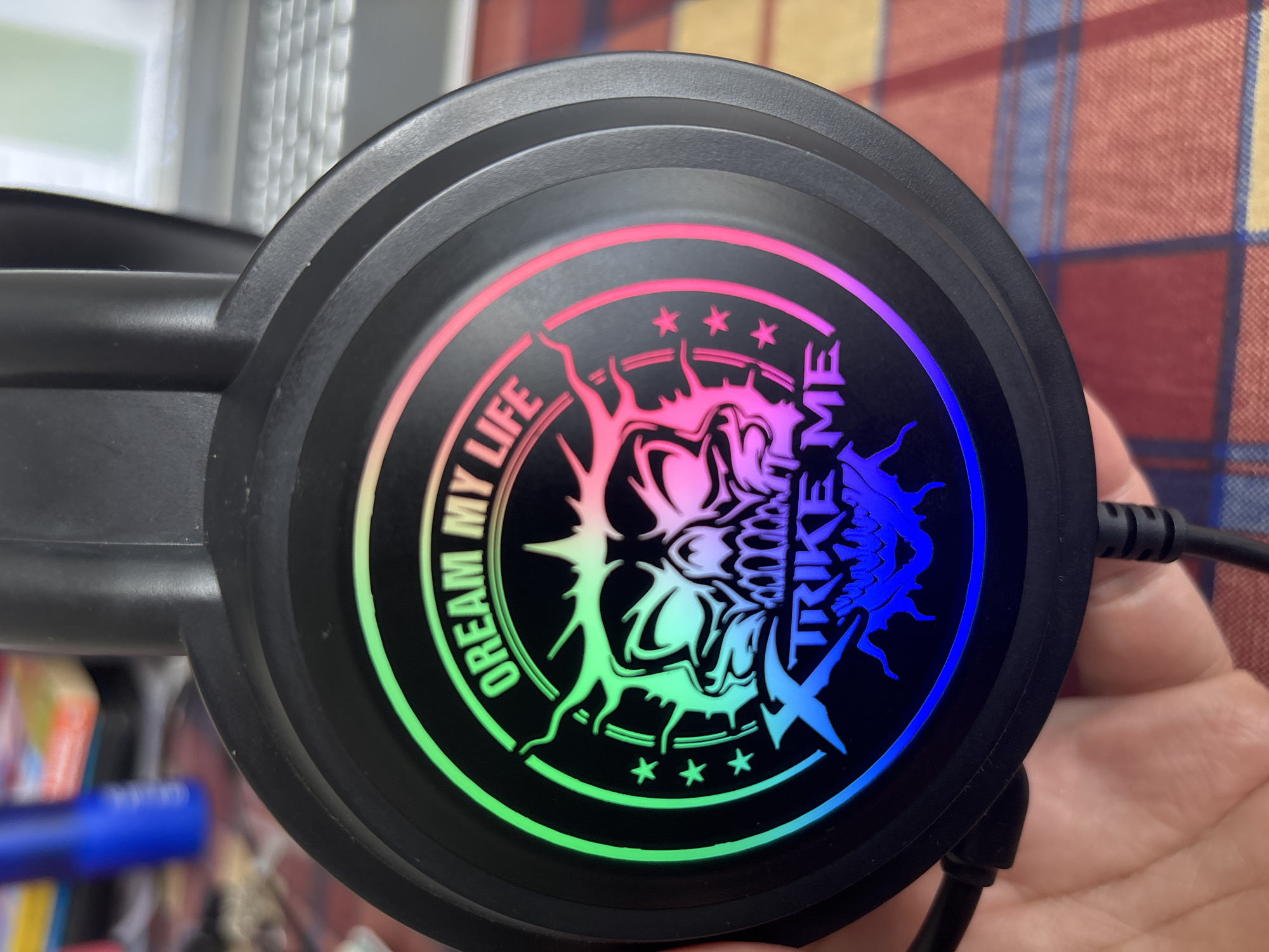 The image shows a close-up of a black headphone earcup with a circular design. The design features a skull with flames, colored in a gradient from green to pink to blue. Text encircles the skull, reading "DREAM MY LIFE" on the left and "TRIKE ME" on the right. Three small stars are visible below the skull. The headphone is being held by a person's hand, which is partially visible. In the background, a plaid tablecloth is visible, suggesting an indoor setting. The lighting appears to be artificial, with the colors of the headphone design being particularly vibrant. No specific time of day, weather, or strong emotions are evident. The image is a product shot, likely intended to showcase the design of the headphones.