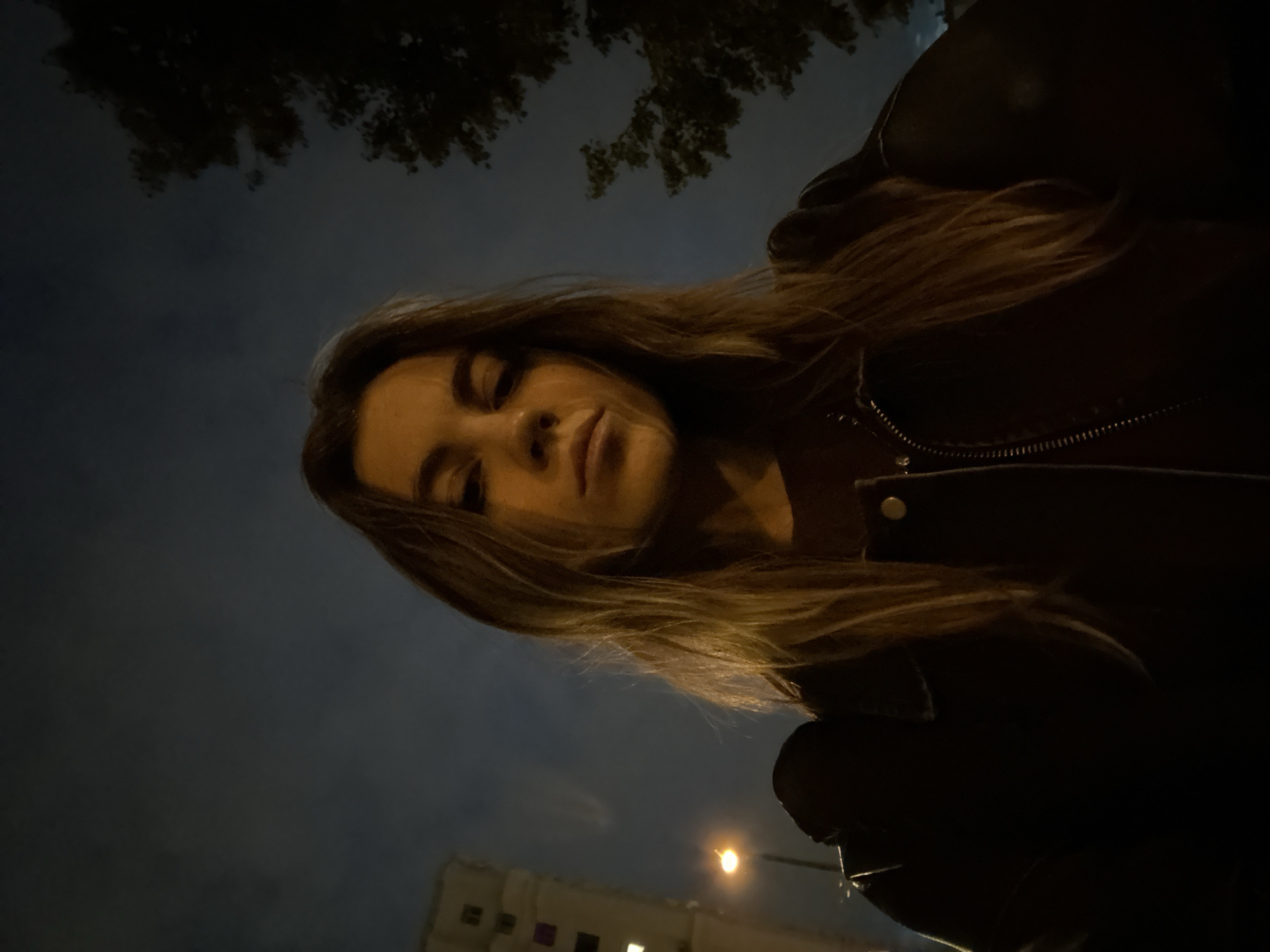 A low-angle, medium shot captures a young woman in a dark setting at night. She has long, light brown hair and is wearing a dark jacket over a brown shirt. Her face is partially illuminated, showing a neutral expression. The background is dark, with a blurry building on the left and trees on the right. A street light is visible to the left of the woman. The overall atmosphere suggests an urban environment at night, possibly Moscow, Russia, given the architectural style of the building, which resembles typical Soviet-era apartment blocks found in the city.