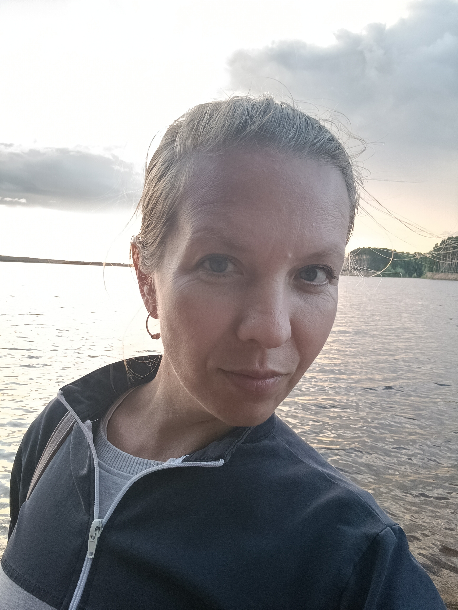 This is a medium-close-up selfie of a woman with blonde hair pulled back, taken outdoors near a body of water. She is wearing a dark gray zipped jacket over a light gray shirt. A thin hoop earring is visible in her right ear. The woman's expression is relaxed, with a slight smile.
The background shows a large body of water, likely a lake or river, with ripples on the surface reflecting the sky. In the distance, a tree line is visible along the horizon, suggesting a natural setting. The sky is partly cloudy, with a bright, diffused light, indicating it is either early morning or late afternoon. The overall mood is serene and calm. The image was taken in Chaikovsky, Russia.