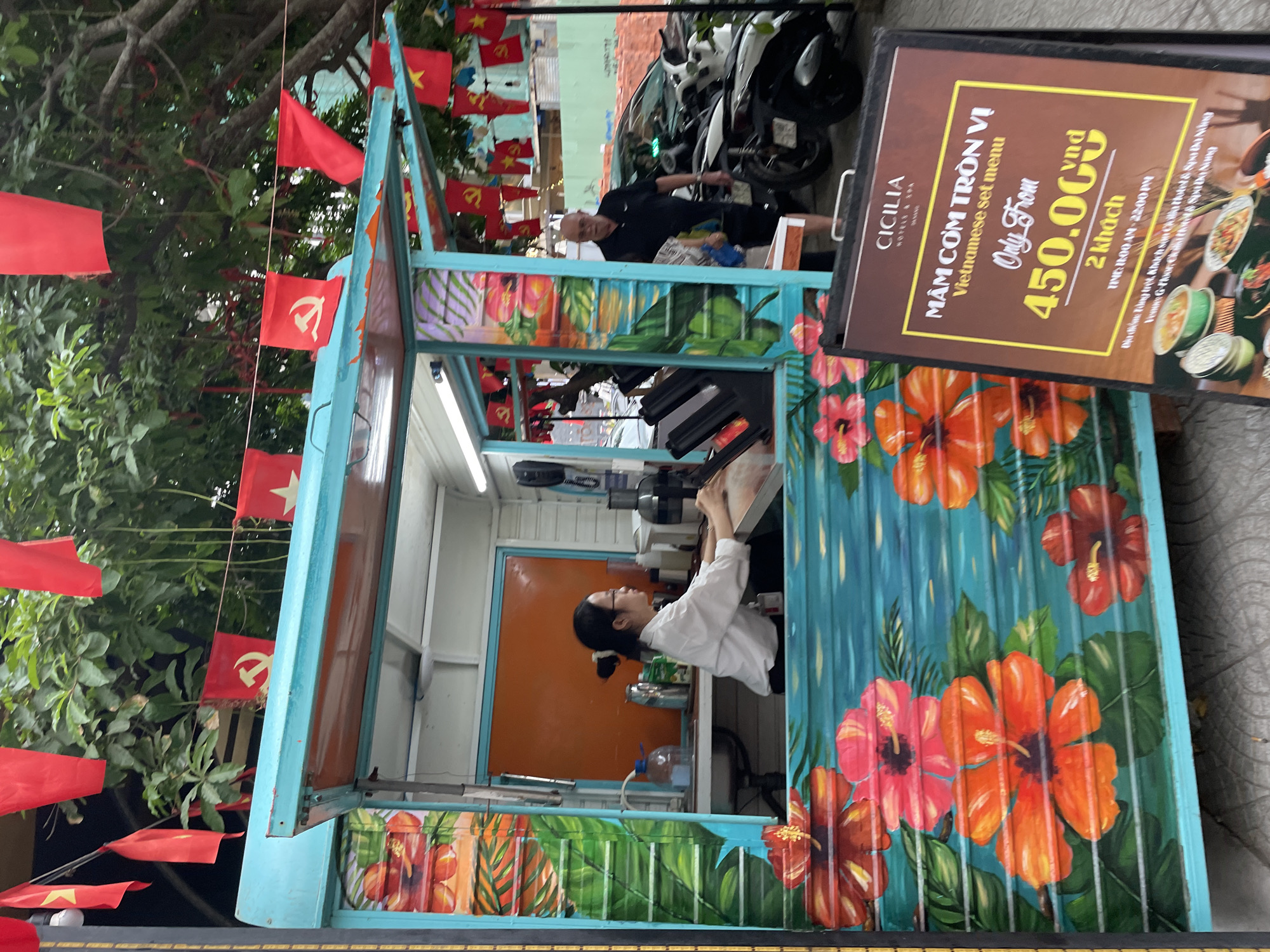 The image shows a food stall in Da Nang, Vietnam, adorned with red flags featuring the hammer and sickle symbol, and yellow stars, suggesting a communist or nationalist theme. A woman in a white shirt is visible inside the stall, possibly preparing food or taking orders. An older man in a black polo shirt stands outside the stall, holding a newspaper. In the background, motorcycles and a car are parked, indicating a street-side location. The stall itself is decorated with colorful floral paintings, primarily hibiscus flowers, against a turquoise background. A sign in front of the stall advertises a "Vietnamese set menu" for 450,000 VND for two people, with operating hours from 11:00 AM to 10:00 PM. The overall scene depicts a vibrant street food setting in Da Nang.