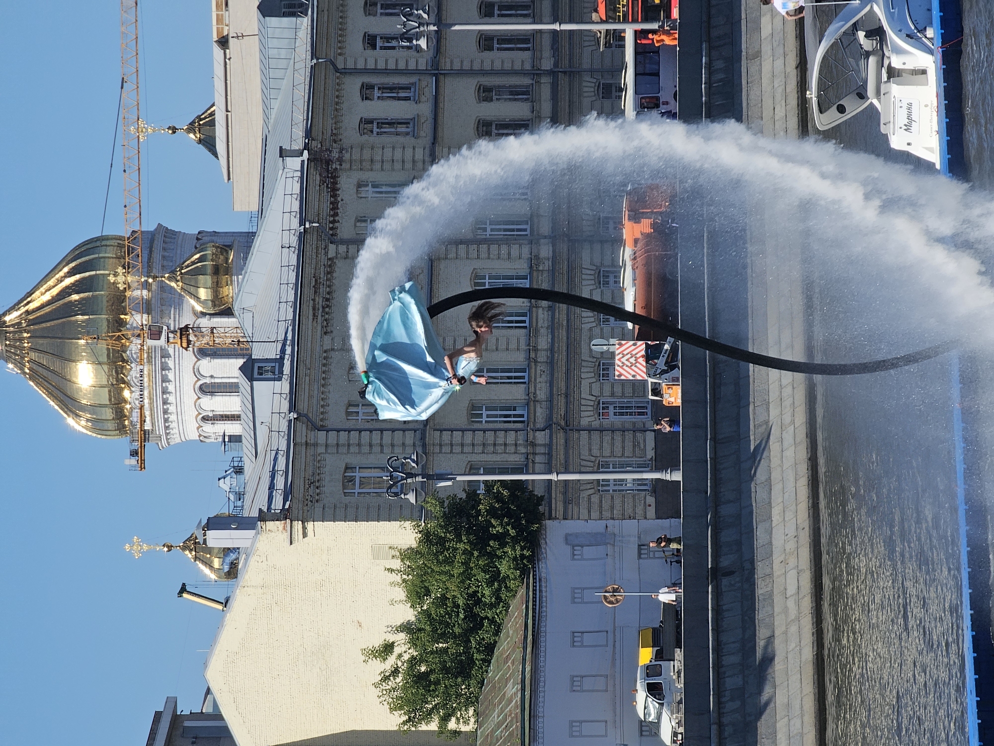 In Moscow, Russia, a person is suspended in the air by a water jetpack over the Moskva River. The individual, wearing a blue fabric garment and seemingly upside down, is the focal point of a performance. Powerful streams of water are directed upwards from a device connected to the performer, creating a dramatic arc of mist and spray that partially obscures the background.
Behind the water spectacle, a wide embankment lines the river, featuring a stone retaining wall. Several vehicles are visible on the embankment, including a white van, an orange tanker truck, and a bus. The architecture of Moscow is evident in the background, with a prominent building featuring ornate detailing and multiple windows. Dominating the skyline are several golden domes topped with crosses, characteristic of Russian Orthodox cathedrals, with construction cranes visible near some of these structures, suggesting ongoing renovation or construction. The sky is a clear, bright blue, indicating fair weather. A white boat with a covered seating area is partially visible in the lower right corner of the frame, with the name "Марина" (Marina) visible on its stern, along with "Санкт-Петербург" (Saint Petersburg).