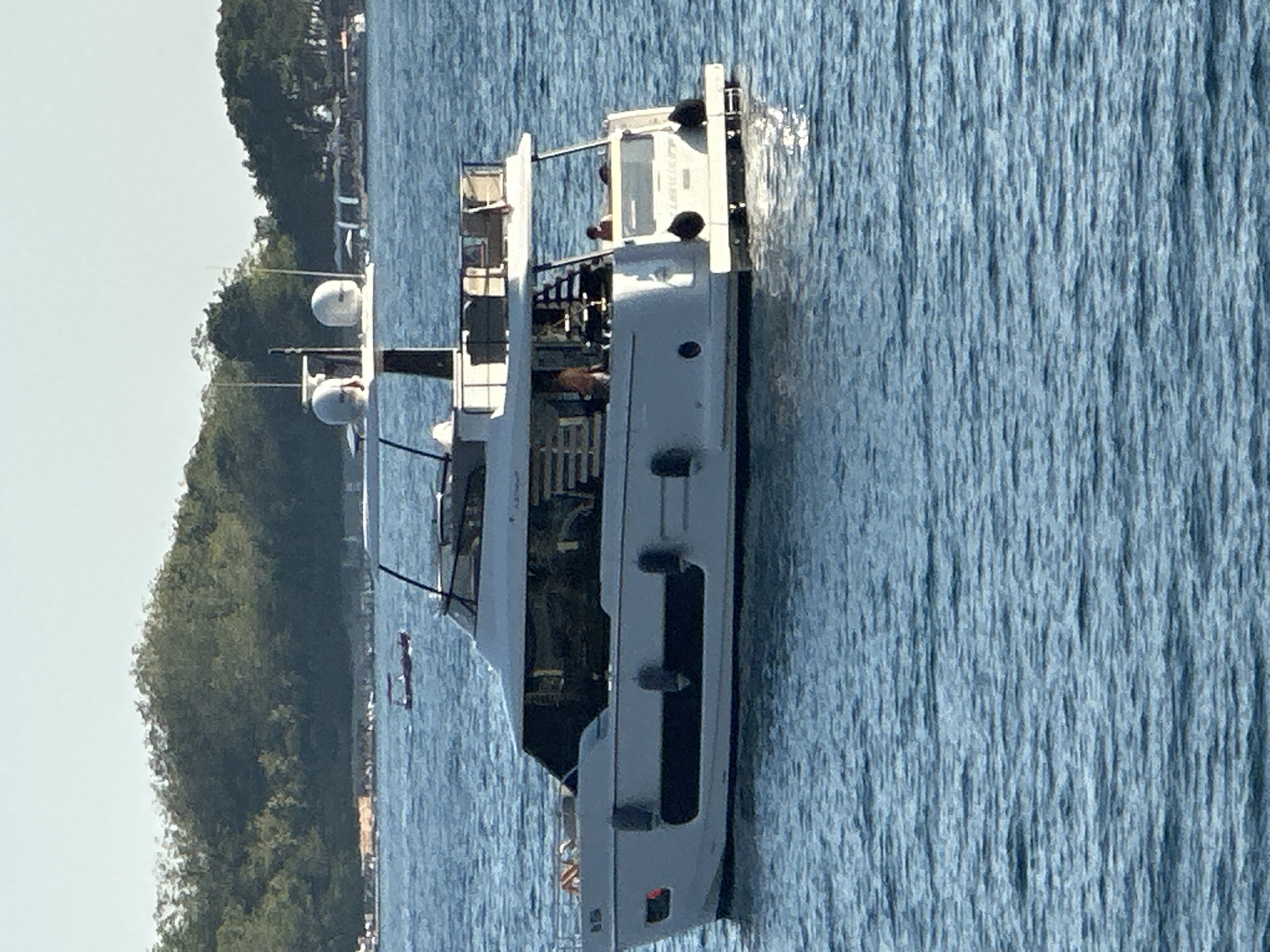 An elevated, long-shot view shows a modern, white motor yacht with multiple levels, anchored in blue water. The yacht has a dark-tinted windowed cabin, a upper deck with seating, and radar domes on its roof. A person is visible on the upper deck, and another on the lower deck. The boat is positioned in front of a lush green, tree-covered hillside, with a hazy, light blue sky above. In the distance, on the left side of the frame, a smaller, red boat with a canopy is also visible on the water. The scene is likely Budva, Montenegro, given the characteristic landscape and coastal setting.