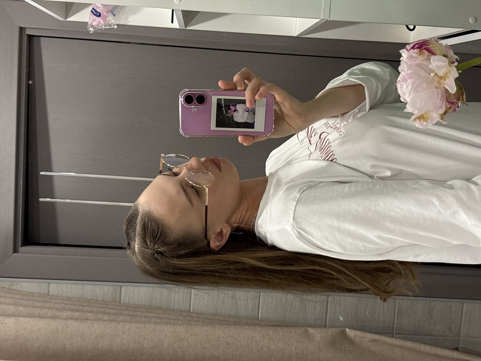 The image is a vertical, close-up shot featuring a young woman in a bathroom setting. She is wearing a white t-shirt and glasses. She is holding up a smartphone in her right hand, and the phone's screen displays a small polaroid photo of two people. A large, delicate pink peony flower is visible on the right side of the frame, partially obscuring her arm. The background includes a dark gray door and a white shelf with a pink object, possibly packaging, on it. The lighting appears to be indoor, and the overall mood is casual and personal. The image is cropped tightly on the woman and the flower, suggesting a focus on personal elements or a self-portrait. The location context is Perm, Russia. No visible text is discernable in the image.