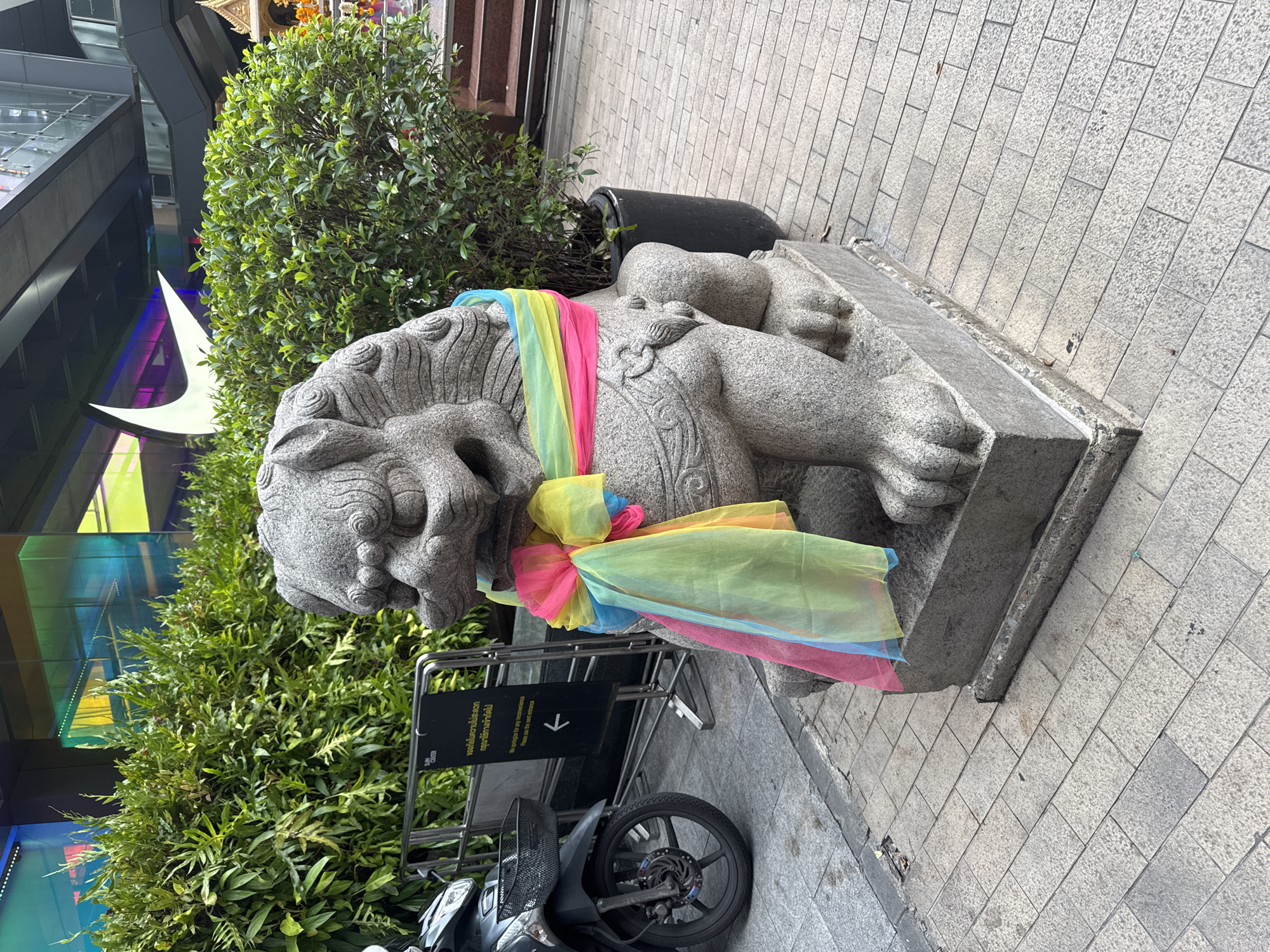 The image shows a stone lion statue adorned with a colorful scarf, likely in Bangkok, Thailand, as the sign in the foreground is written in both Thai and English. The statue is resting on a stone base, positioned beside a planter containing shrubbery. A black sign with white text is visible near the statue, along with the front wheel of a parked motorbike. The background includes greenery and a building with modern architectural elements. The pavement is made of grey rectangular tiles.